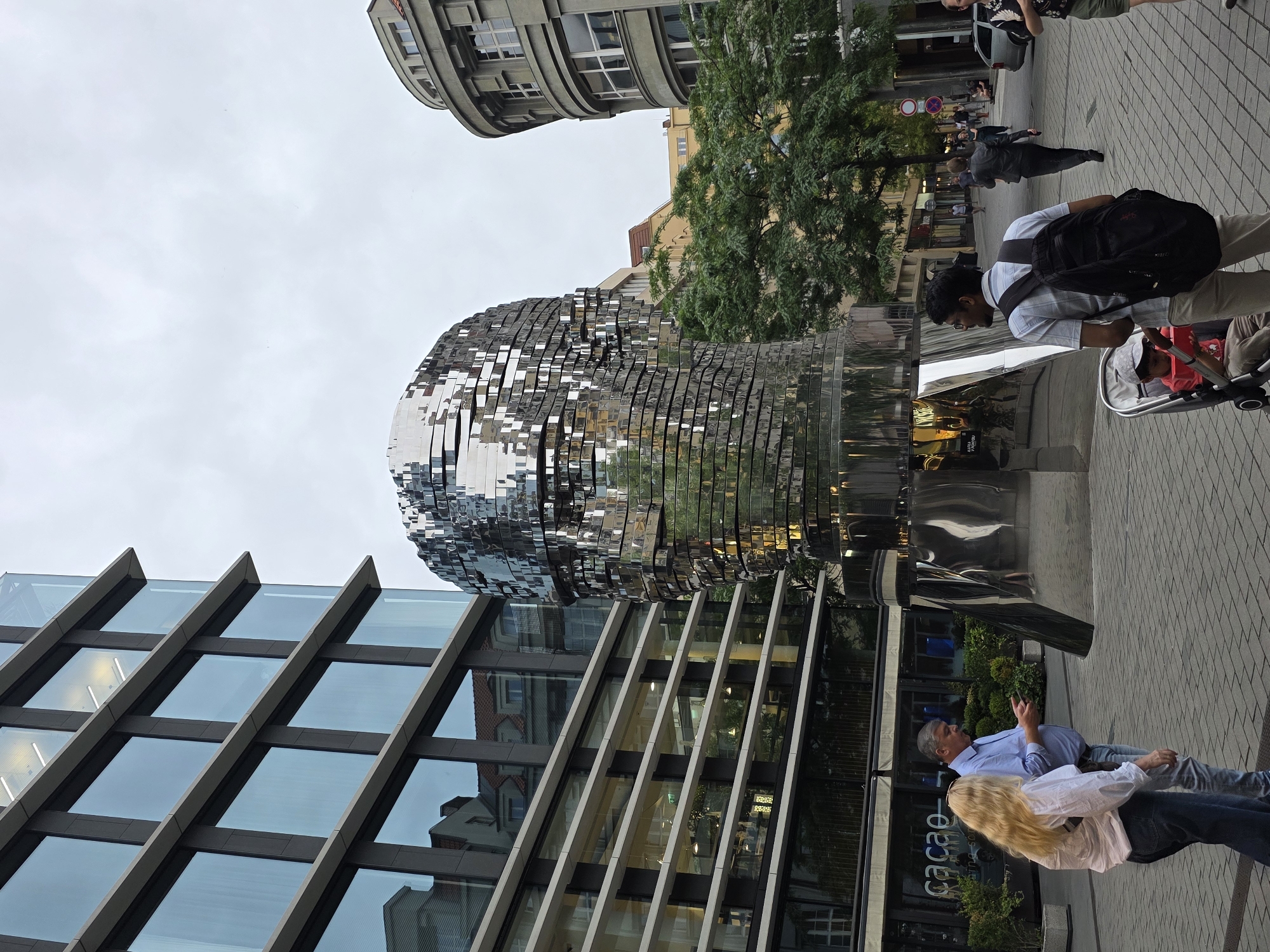 An urban street scene unfolds under an overcast sky, prominently featuring the "Head of Franz Kafka" sculpture by David Černý. The large, reflective, kinetic sculpture, composed of numerous horizontal, layered metallic segments, dominates the right side of the frame, appearing as a massive, abstract head that reflects its surroundings.
To the left, a modern building with a glass facade and diagonal structural beams stands tall. In the background, behind the sculpture and some green trees, an older, curved building with intricate detailing and numerous windows, possibly in an Art Nouveau style, adds architectural diversity to the street.
The paved sidewalk is populated with several pedestrians. In the mid-ground, a man wearing a light shirt, dark trousers, and a black backpack is seen pushing a baby stroller, in which a child in a red top is seated. Closer to the foreground, a man with gray hair, dressed in a blue shirt, stands and gestures while seemingly conversing with a blonde woman. Other people are visible walking in the background.
The sky is a bright, uniform gray, indicating an overcast day. Lush green foliage from trees adds natural elements to the urban landscape. On the ground level of a building in the foreground, a storefront sign is partially visible, showing the words "cacao" and "pralines chocolaterie," suggesting a chocolate shop or cafe. The scene captures a typical bustling daytime environment in a city, characterized by a mix of historic and contemporary architecture.