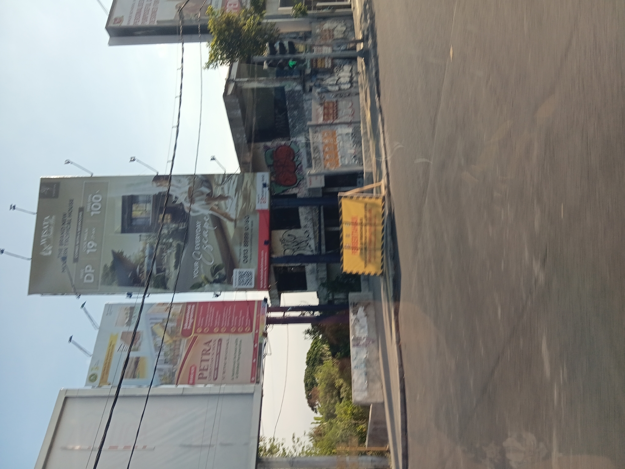 This is a medium shot of a street scene in Surabaya, Indonesia, taken on a clear, sunny day. The image features several large billboards, a building with graffiti, a traffic light, and a road. The main billboard on the right advertises "WISATA" and a "BRAND NEW TROPICAL HOUSE" with images of a house and people. Another billboard on the left promotes "PETRA CHRISTIAN SCHOOL." The building in the background is covered in graffiti, with a traffic light showing a green signal on the right. The foreground shows a paved road with some faint markings. The overall environment appears urban, with a sense of commercial activity.