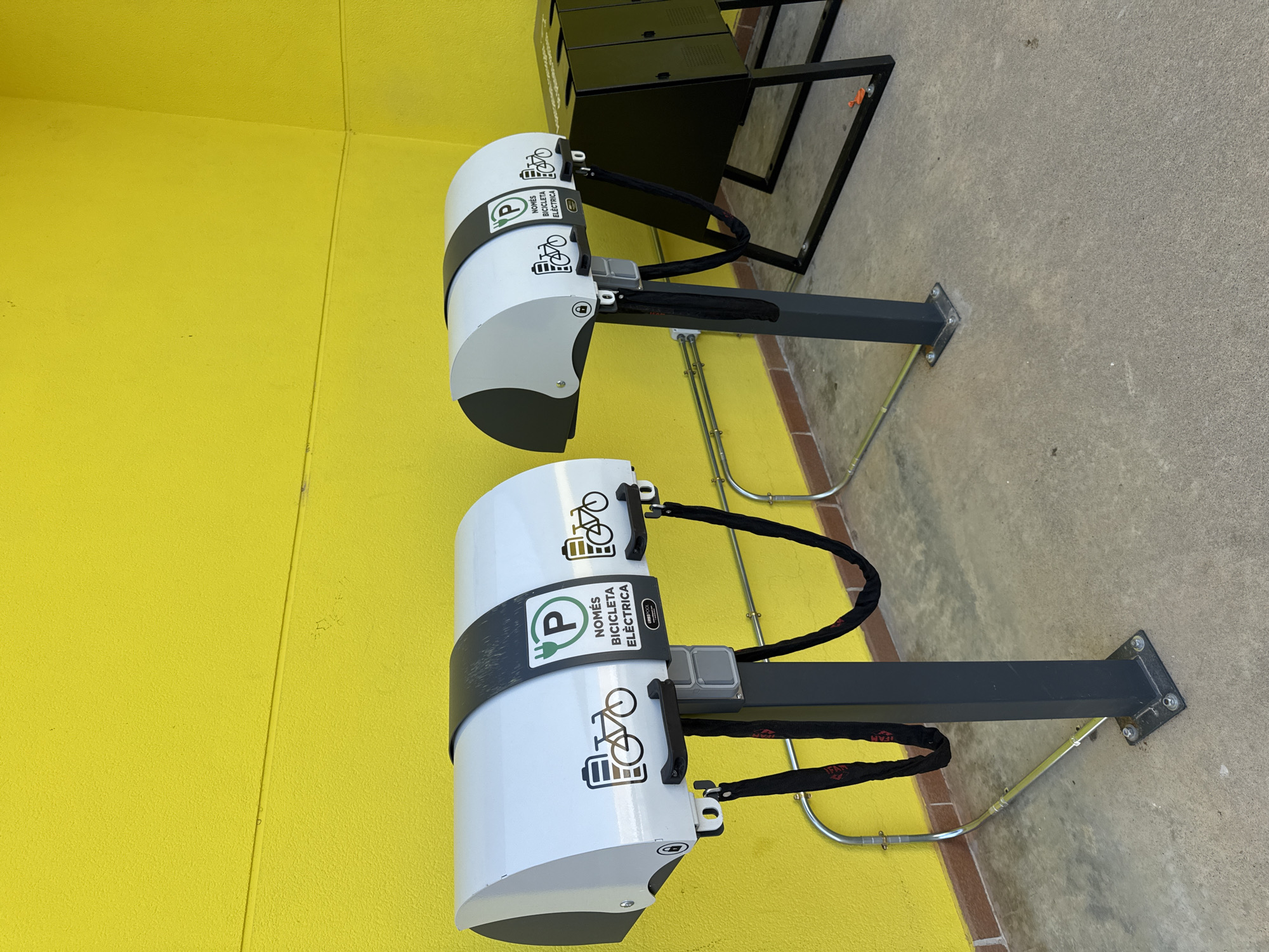 The image shows two electric bicycle charging stations positioned in front of a bright yellow textured wall. Each station has a white, curved housing with a black stripe around the top. The housing features icons of a bicycle and a battery, along with text that appears to be in Catalan: "NOMÉS BICICLETA ELÈCTRICA" and "P" inside a green circle, indicating parking for electric bicycles. A black chain hangs from each station, presumably for securing the bicycles. To the right of the charging stations is a black metal rack with multiple compartments. The ground is concrete, and metal conduits run along the wall at the base. The scene appears to be an outdoor public space, possibly a street or a park, in the city of Vidreres, Spain, given the Catalan text. No people are visible in the image.