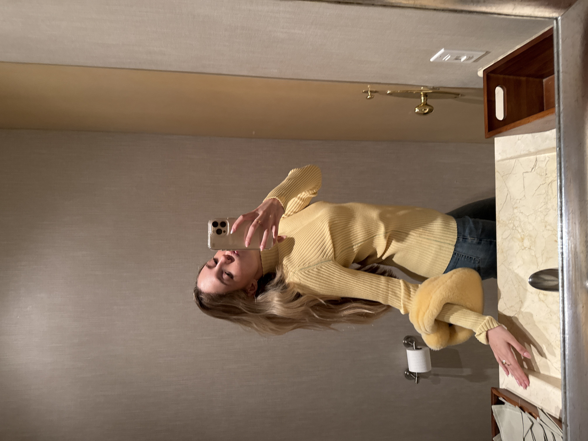 A woman is taking a selfie in a bathroom mirror. She is wearing a yellow sweater and blue jeans. The bathroom has light-colored walls and a marble countertop. There is a gold faucet and a wooden shelf on the wall. The image was taken in Mexico City, Mexico.