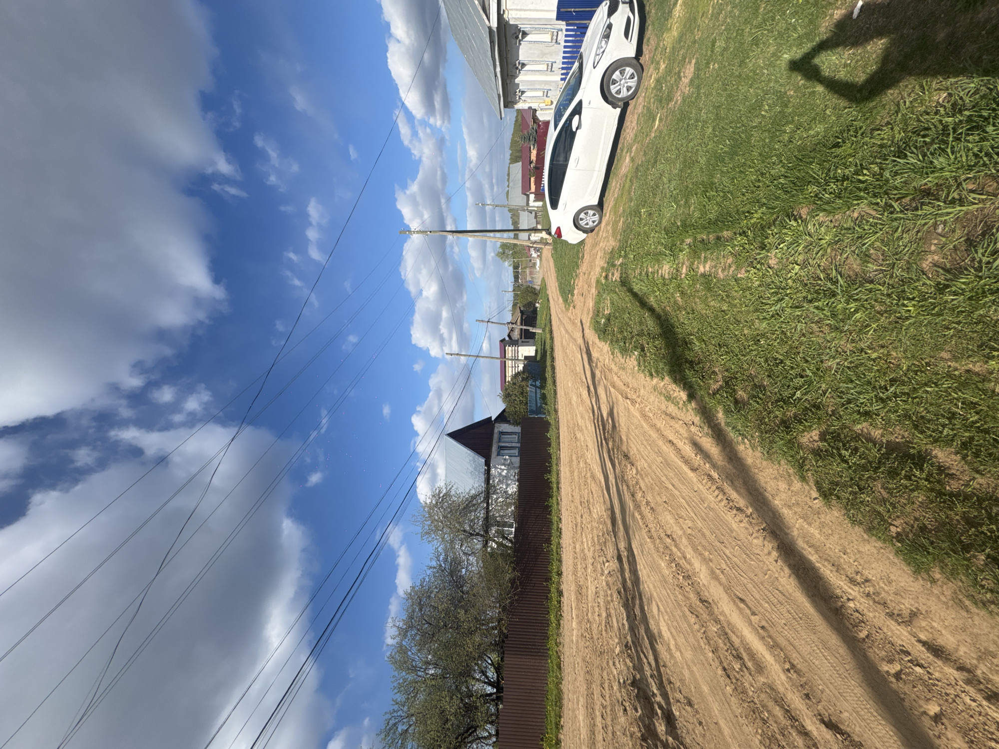 The image depicts a rural scene in Ol'khovochka, Russia. A dirt road runs through the center, flanked by grassy areas. A white car is parked on the right side of the road. A brown fence and houses line the left side, with utility poles and power lines visible overhead. The sky is blue with scattered clouds. The shadow of the person taking the photo is visible on the right side of the road.