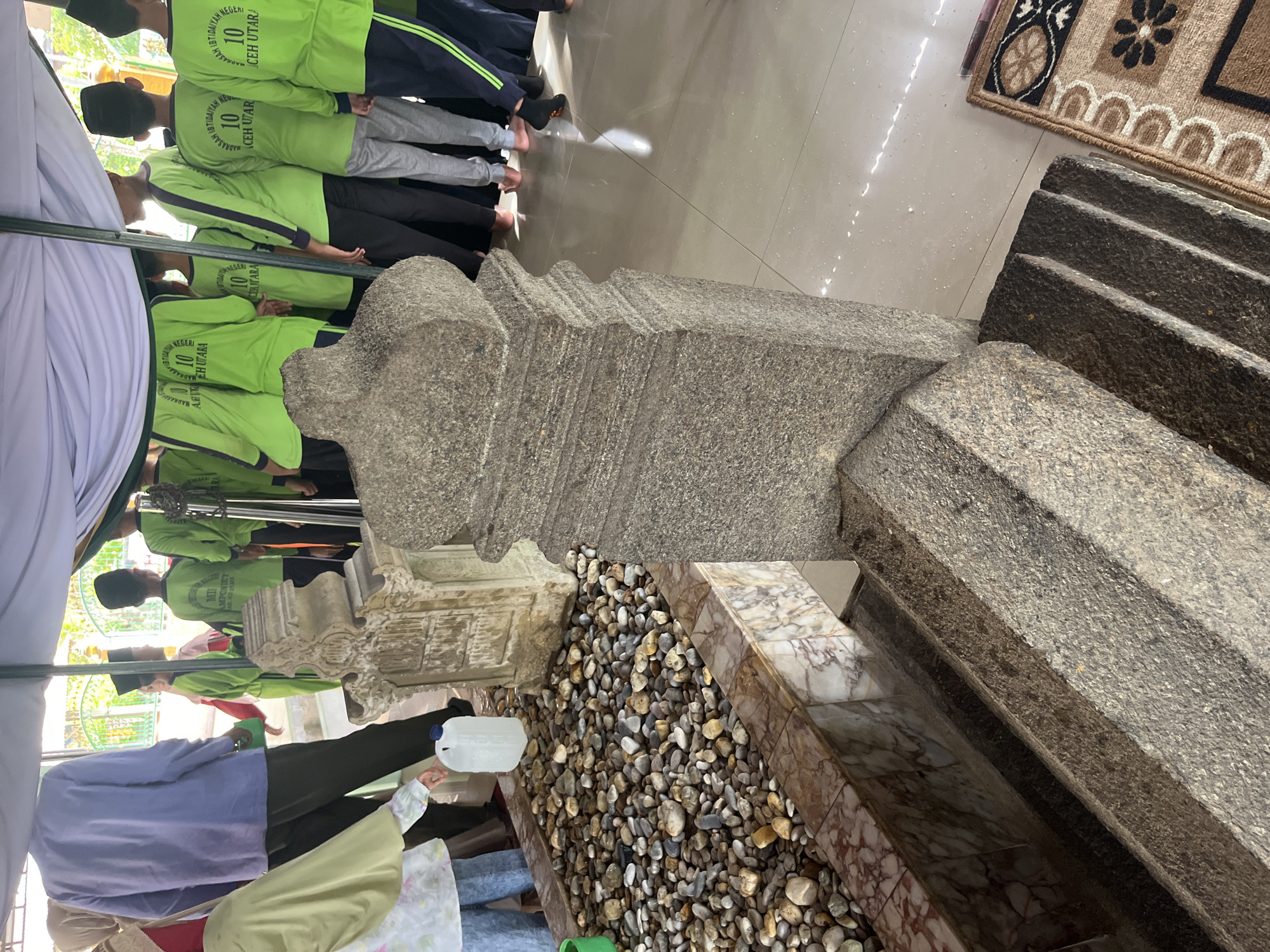 In Beuringen, Indonesia, an outdoor scene is captured, likely a historical or religious site. The foreground features a stone tomb with steps leading up to it, constructed from rough-hewn granite. Beside the tomb is a grave enclosure filled with pebbles and a white plastic jug with a blue cap. In the mid-ground, a group of children, some wearing light green shirts with the text "MADRASAH IBTIDAIYAH NEGERI 10 ACEH UTARA" and black skullcaps, stand under a white canopy. Some of the children are wearing black pants, while others are in light grey pants. To the left of the tomb, an adult in a light blue long-sleeved shirt and dark pants stands. The ground is a tiled floor that reflects the light. A small rug with a patterned design is visible on the right side of the frame.