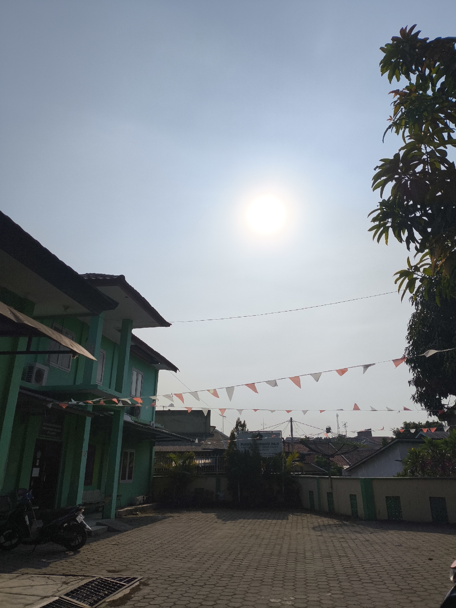 In Cilegon, Indonesia, a two-story building, painted green with white trim, stands on the left side of the image, its dark roof extending outwards. The building has multiple windows and an air conditioning unit is visible. The foreground is paved with interlocking grey bricks. Two motorcycles are parked in front of the building. Strings of red and white triangular flags are strung across the scene, above a low wall. The sky is bright, with the sun prominently visible and emitting a strong glare. A large tree with dark green leaves is on the right side of the image. In the background, other buildings and rooftops can be seen, along with satellite dishes and antennas. A sign is visible on a bush in the mid-ground which reads "Pusat Informasi Haji KEMENAG KOTA CILEGON".