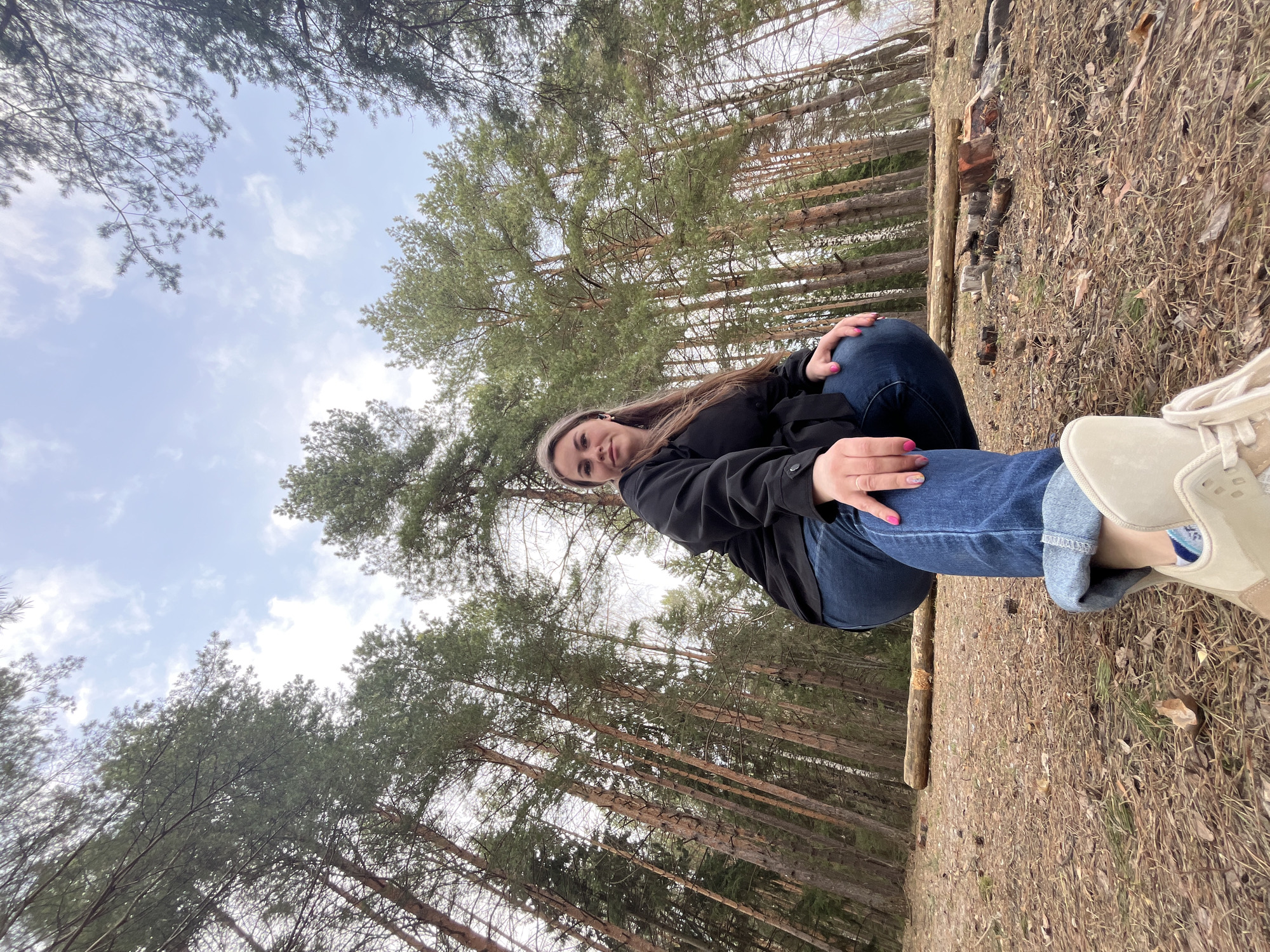 The image shows a woman sitting on a log in a forest. She is wearing a black jacket and blue jeans, with a headband in her hair. Pine trees are visible in the background. The location is inferred to be in the city of Chaikovsky, Russia based on prior context. The ground is covered with dry grass and pine needles. A small pile of burned wood is visible near the log. The sky is visible through the trees, with some clouds present.