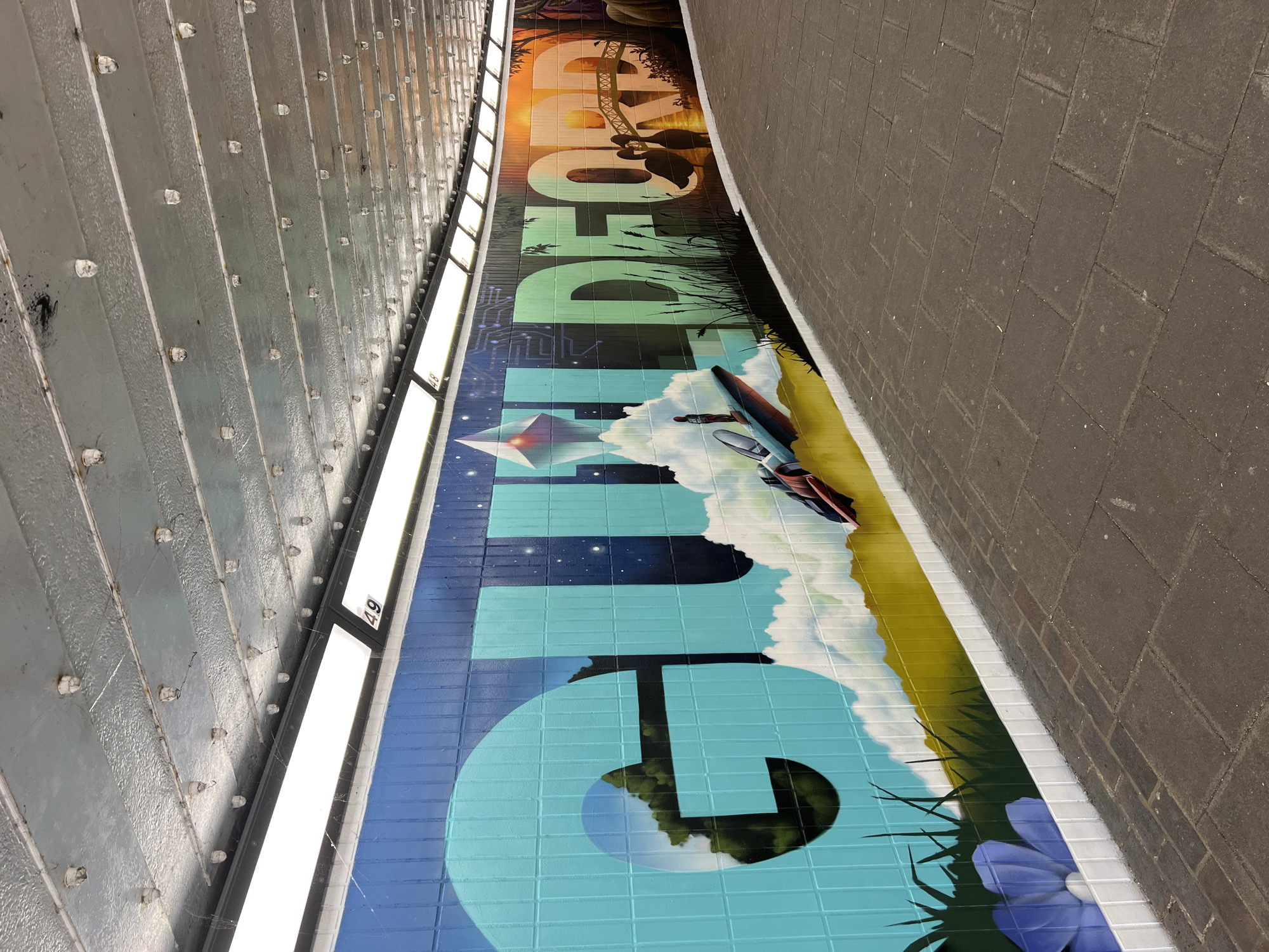 The image shows a section of a mural painted on a curved wall in a pedestrian underpass. The mural features large, stylized lettering spelling out "GUILDFORD". The letters are filled with various scenes and imagery. The "G" and parts of the "U" and "I" are filled with a green forest and a blue sky with clouds and stars. A futuristic spacecraft with a pilot is depicted in front of these letters, alongside a geometric, crystal-like structure. The letters "L", "D", "E", "F", "O", "R", and "D" transition from green to a warm orange and yellow sunset. This section of the mural includes silhouetted figures of two swans on a bridge, and trees and possibly elephants in the background. The wall itself is tiled with rectangular bricks, and the mural is painted over these tiles. The underpass has a corrugated metal ceiling with rows of lights and some visible cobwebs. The ground in the underpass is paved with dark rectangular bricks. The scene appears to be located in the city of Guildford, United Kingdom, as indicated by the mural's text.