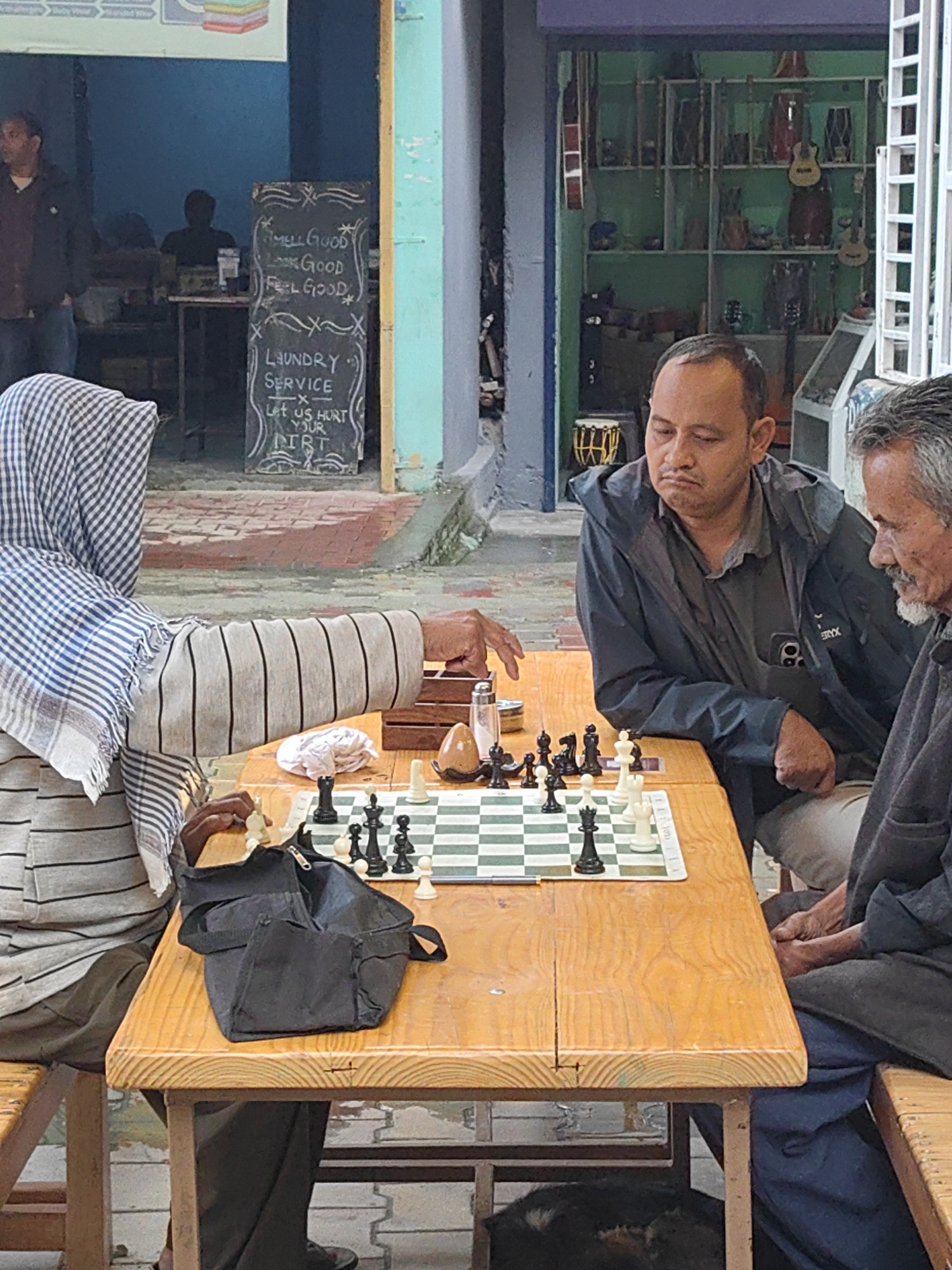 In Vashisht, India, three individuals are gathered around a wooden table, engaged in a game of chess. Two men are seated on one side, facing a third person who is largely obscured by a checkered headscarf and striped shirt. The person in the headscarf is actively playing, with their hand reaching for a white chess piece. The man in the dark jacket closest to the viewer watches the game with a pensive expression, while the third man, older with graying hair, appears to be observing the game with a slight smile.
The chessboard, positioned centrally on the table, displays an active game in progress. Surrounding the board are various items: a salt shaker, a small container, a white cloth, and a brown egg-shaped object. A black bag rests on the table near the playing individual.
The environment suggests an outdoor setting, possibly a street or a small shop's exterior, indicated by the brickwork visible in the foreground and the building facades in the background. One building features a chalkboard sign with handwritten text that reads: "SMELL GOOD LOOK GOOD FEEL GOOD. LAUNDRY SERVICE. LET US HURT YOUR DIRT." Another building in the background displays shelves filled with musical instruments, including guitars and drums. The overall scene captures a moment of leisure and social interaction in a bustling Indian locality.