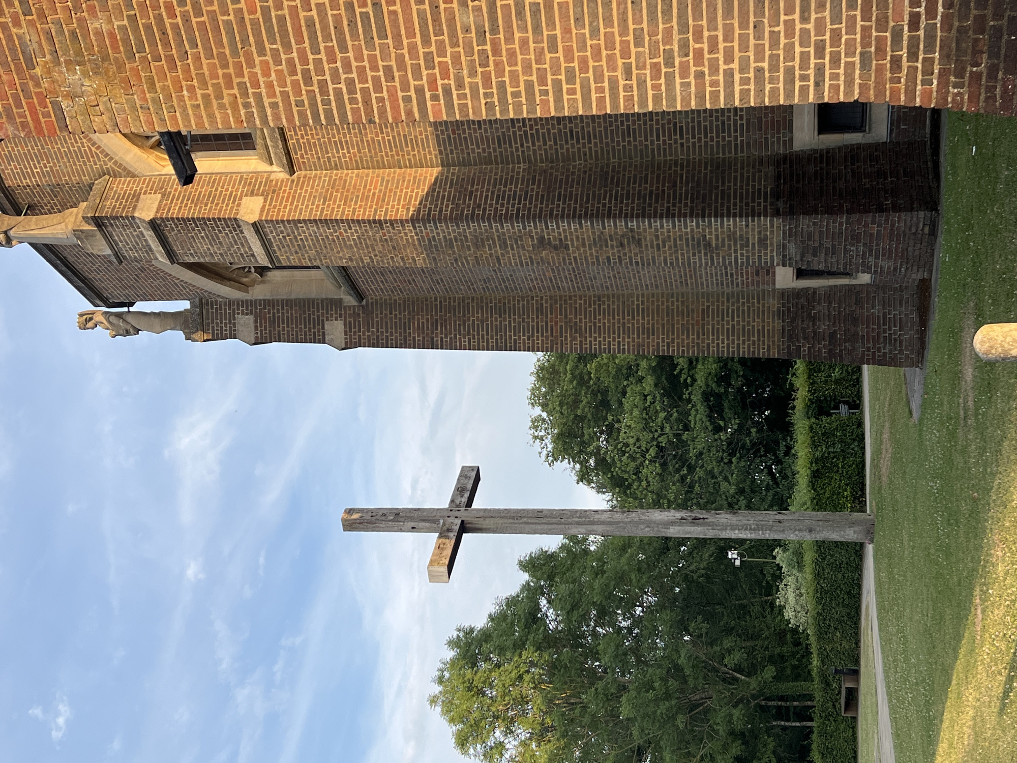 This is an outdoor photograph taken in Guildford, United Kingdom. The image features a large wooden cross standing prominently in the foreground. Behind the cross, a section of a brick building, possibly a church, is visible. The building has a textured brick facade and a sloped roof. To the right of the cross, a manicured green lawn stretches out, bordered by a neatly trimmed hedge and lush trees. The sky above is a clear blue with wispy white clouds, suggesting fair weather during the daytime. The lighting indicates it is likely late afternoon, with soft sunlight illuminating the scene and casting gentle shadows. There is no visible text in the image. The overall atmosphere is peaceful and serene, with the prominent cross suggesting a religious or memorial context.