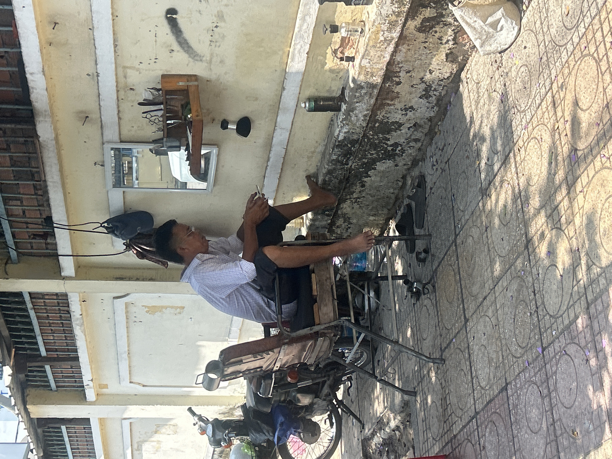 A Vietnamese man sits in a barber chair mounted on a motorcycle on a sidewalk in Nha Trang, Vietnam. He is wearing a blue and white checkered shirt and black shorts, holding a phone in his right hand. The scene is set against a peeling, dirty wall with a mirror and a small wooden rack holding barber tools. Various canisters and bottles sit on a ledge. The sidewalk is tiled with patterned gray stones, and purple flower petals are scattered across the ground. Sunlight casts shadows on the sidewalk.