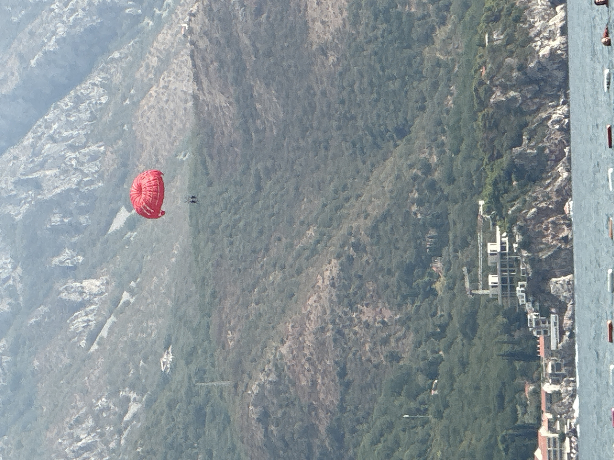 In a wide shot of Boreti, Montenegro, two figures are parasailing over a body of water, with a red parachute aloft. The parasailers are suspended in the air, with the parachute acting as a sail. In the background, a mountainous terrain is covered in dense vegetation, with a few scattered buildings. A construction site is visible on the side of a hill, with cranes and unfinished structures. The water in the foreground is choppy, with several buoys floating on the surface.
