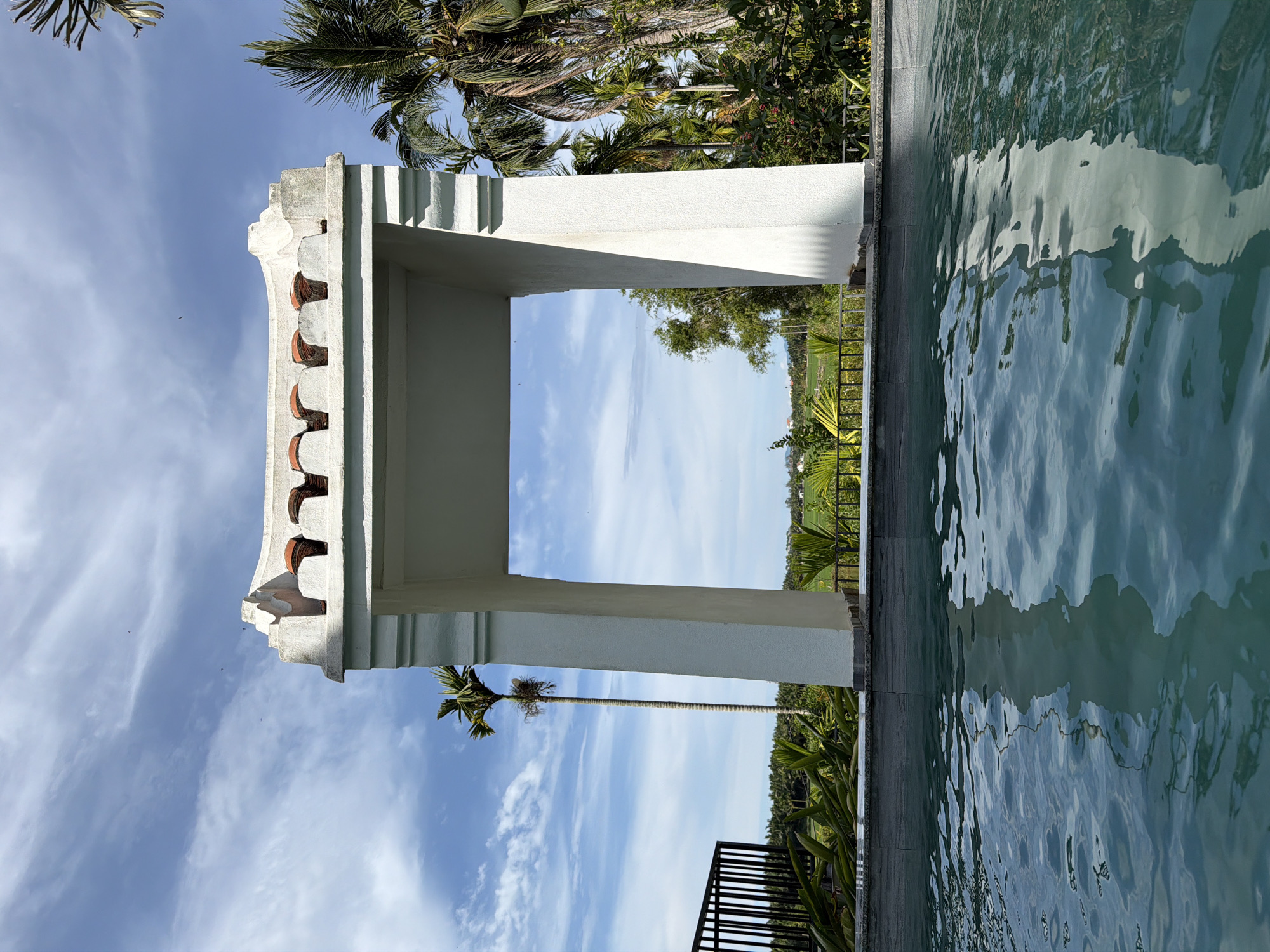 A white, arched gateway with a decorative, segmented roof stands in the foreground, framing a view of a tropical landscape under a blue, cloudy sky. The gateway is made of concrete and features geometric detailing. In front of the gateway is a swimming pool with clear blue water, reflecting the gateway and the sky. Beyond the pool and gateway, lush greenery, palm trees, and a distant field of crops are visible. The scene suggests a tranquil resort or private residence in a tropical setting. This location is likely in the countryside surrounding Hoi An, Vietnam, a region known for its picturesque rice paddies and tropical foliage. There are no people present in the image, and the environment is calm and serene.