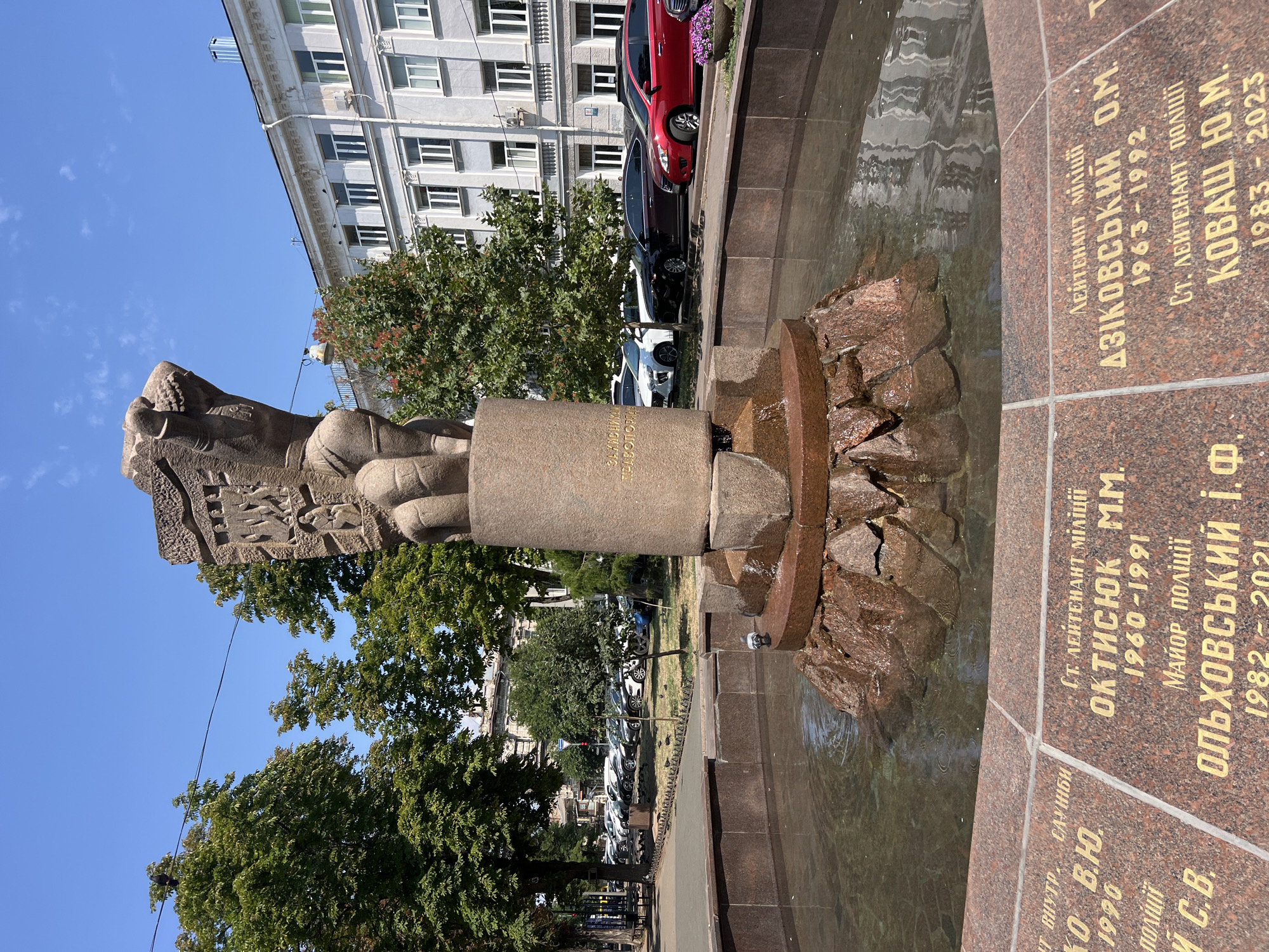 A low-angle, medium shot captures a stone sculpture and fountain in Odesa, Ukraine, on a clear, sunny day. The sculpture, made of a rough-textured, reddish-brown stone, depicts a figure with a carved headdress, a bent torso, and legs resting on a cylindrical base. The base has faint, golden Cyrillic inscriptions on its side. Water cascades from the sculpture into a circular fountain pool, which is lined with similar reddish-brown stone. Around the fountain's edge are engraved granite plaques bearing golden Cyrillic lettering and dates, presumably memorializing individuals. In the background, several mature green trees frame the scene, with the facade of a light-colored, multi-story building visible behind them. A row of parked cars, including a bright red sedan, is seen along the street in front of the building. Pigeons are scattered around the fountain and on the ground. The sky is a vibrant blue with a few wisps of white clouds.