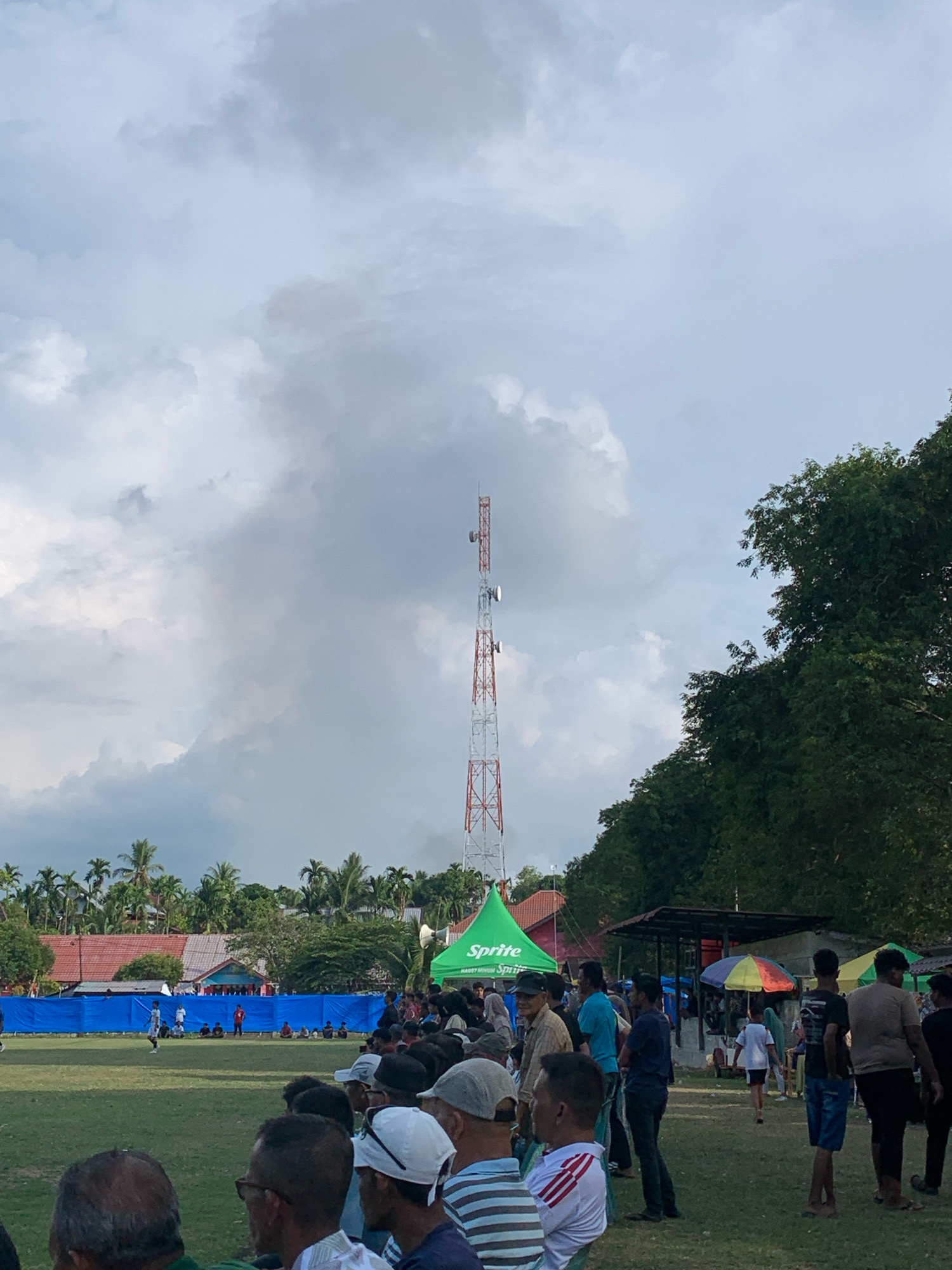 The image depicts a crowd gathered for an outdoor event, likely a soccer match, on a grassy field in Keudelapang, Indonesia. A communication tower stands tall in the background against a cloudy sky. In the mid-ground, a blue barrier separates the field from spectators, and tents are set up, including one branded with "Sprite." People of various ages are visible, some watching the event, others milling about. The environment includes trees and buildings, characteristic of a semi-urban or rural setting.