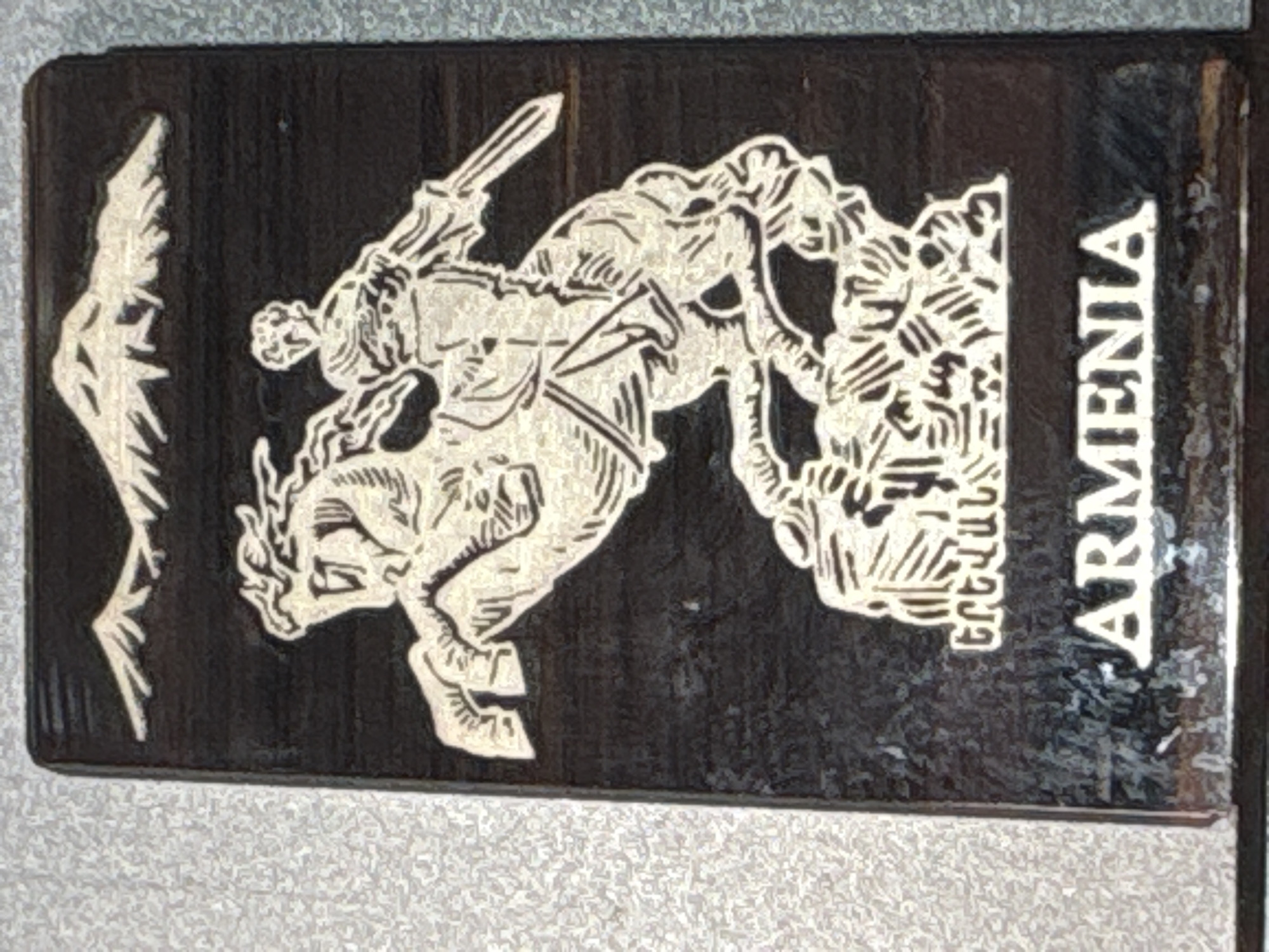 The image displays a rectangular, dark-colored item, likely a money clip or decorative plaque, featuring a prominent two-tone engraving. The central subject is a heroic warrior on horseback, depicted in a dynamic pose, rearing up as he wields a sword over his head. He is engaged in combat with a multi-headed or complex serpentine creature or dragon positioned on the right side of the composition. A stylized wing-like element is visible on the far left.
The engraving is rendered in a light, possibly white or cream color, contrasting sharply with the dark, almost black background. The background appears to have a subtle texture, possibly resembling wood grain or brushed metal. Along the right edge of the item, vertical text is engraved. The word "ARMENIA" is clearly visible in large, uppercase Latin letters. Directly beneath this, in smaller Armenian script, the word "ԵՐԵՎԱՆ" (Yerevan) is etched.
The scene strongly evokes the legend of Saint George and the Dragon, a powerful symbol of victory and heroism often found in Armenian iconography. The item's design, combining this imagery with the country and city names, suggests a commemorative or souvenir piece. This item was photographed in Krasnoyarsk, Russia.