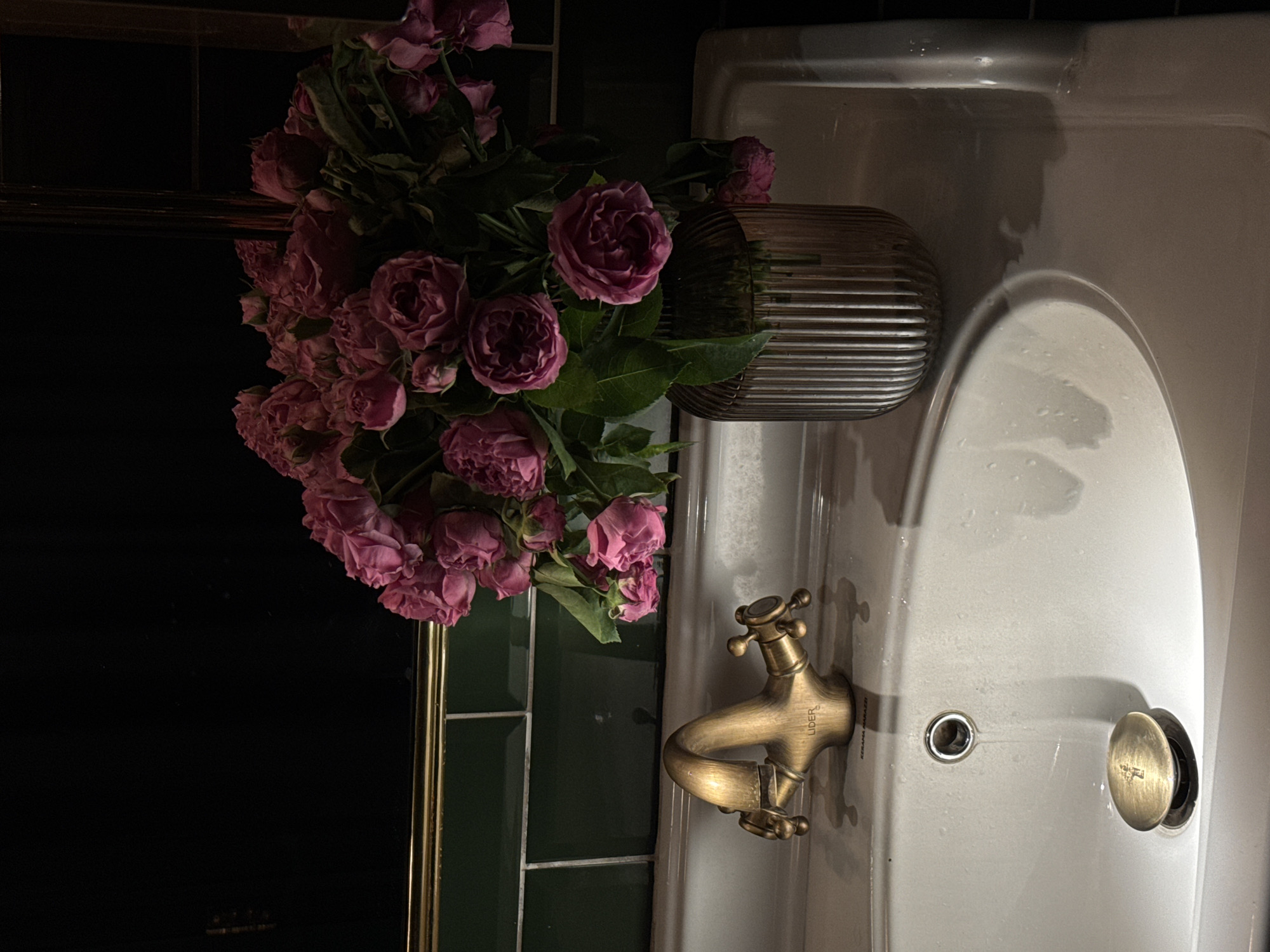 A top-down shot reveals a sink area inside a restroom. A bunch of purple flowers sits in a ridged silver vase next to the sink. The faucet is brass and labeled "Lider." The drain is visible on the right side of the sink. The wall behind the sink is tiled in dark green and black. There's no visible information to confirm this is in Yerevan, Armenia.