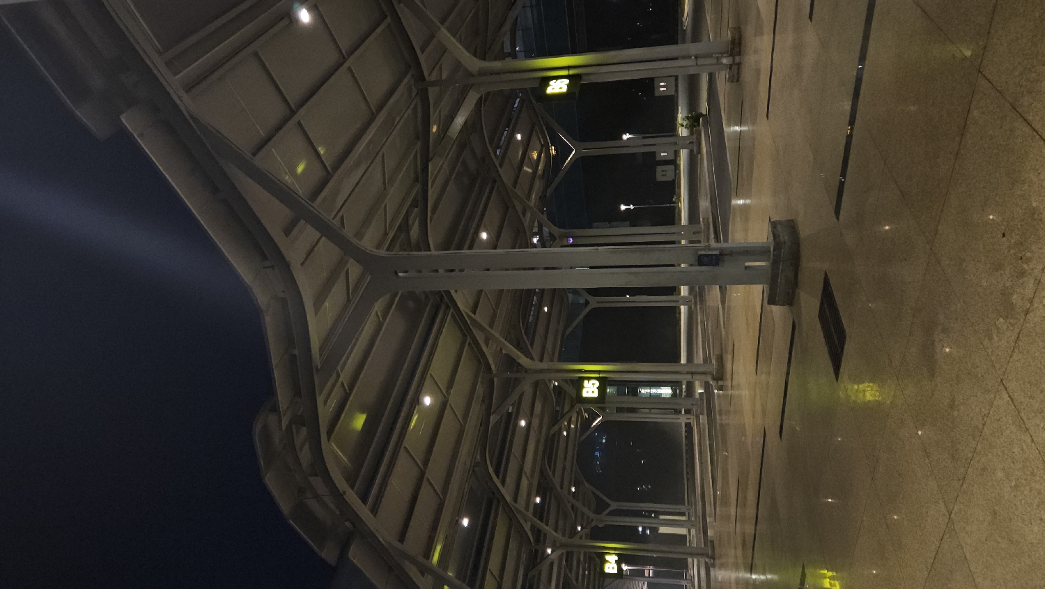 The image depicts an illuminated modern architectural space at night, likely an airport terminal or a similar large public building, situated in Cam Ranh, Vietnam. The scene showcases a covered area with a distinctive structural design.
The primary subject is a series of robust, light-colored columns or beams that support an overhead canopy or roof. These structural elements branch upwards and outwards, creating a repeating, geometric pattern against the dark sky visible in the background. The ceiling or underside of the canopy is paneled and contains numerous small, embedded circular lights that cast a warm glow.
The floor is a smooth, reflective tiled surface, possibly polished stone or marble, with dark linear strips forming a grid pattern. Reflections of the overhead lights are visible on this glossy surface. Beyond the covered structure, the dark expanse of the night sky is visible, with faint, blurry artificial lights in the distance, indicating an outdoor or semi-outdoor setting.
A notable detail is the presence of several bright, glowing yellow-green rectangular signs. One clearly displays "B5", and another partially visible sign beneath it shows "B4". These signs likely indicate specific gates or sections within the facility. No people are visible in the image, contributing to a serene and perhaps deserted atmosphere. The overall impression is one of modern infrastructure designed for efficiency and nighttime functionality.