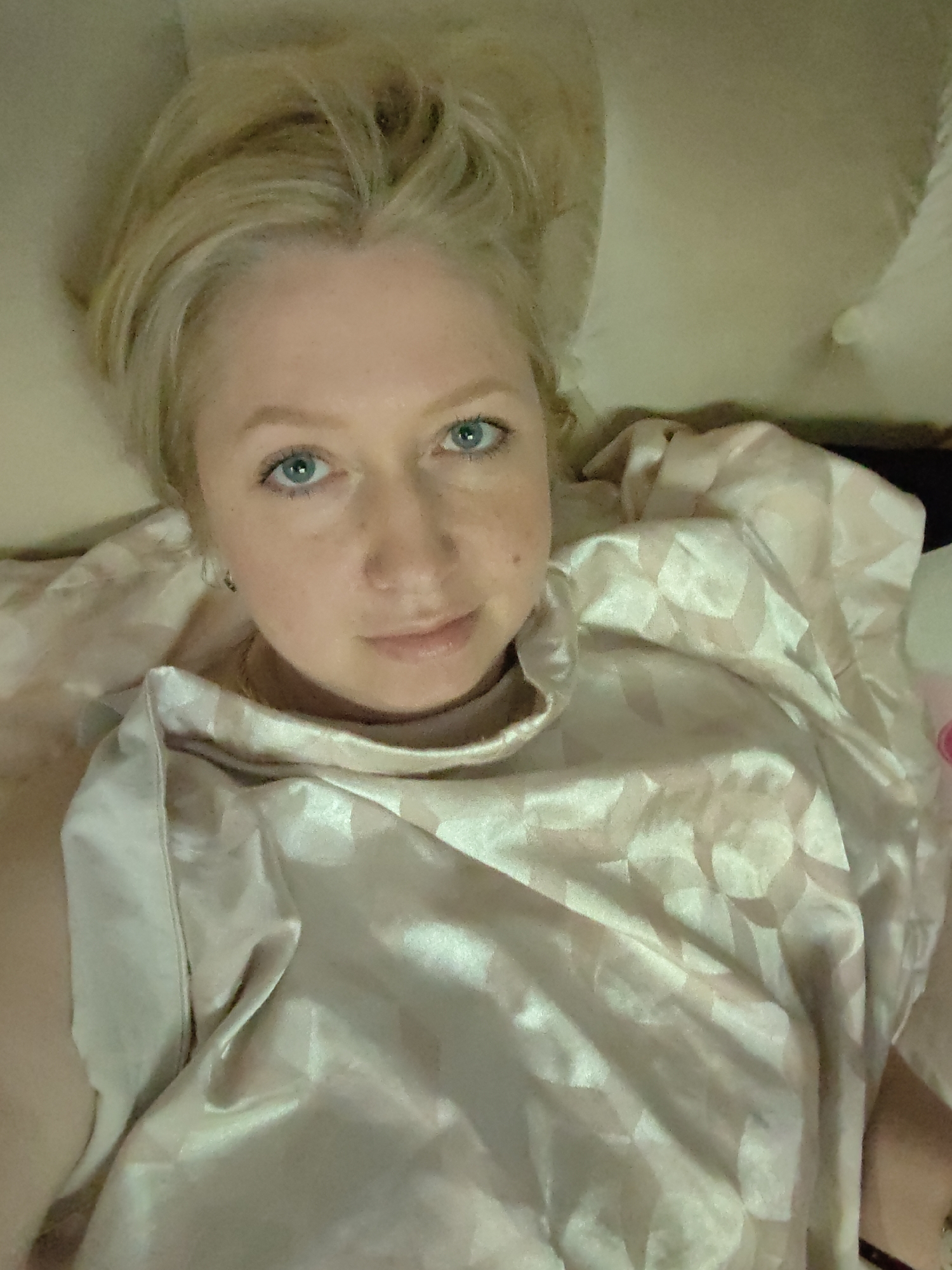 The image shows a woman lying on her back, facing the camera, with her head resting on a light-colored surface. She has blonde hair and blue eyes. She is wearing a light-colored, silky garment with a subtle pattern. The background is out of focus and appears to be indoors. There is no indication of the location being the city of Sochi, Russia, or any other specific location. The scene is a close-up portrait of the woman.