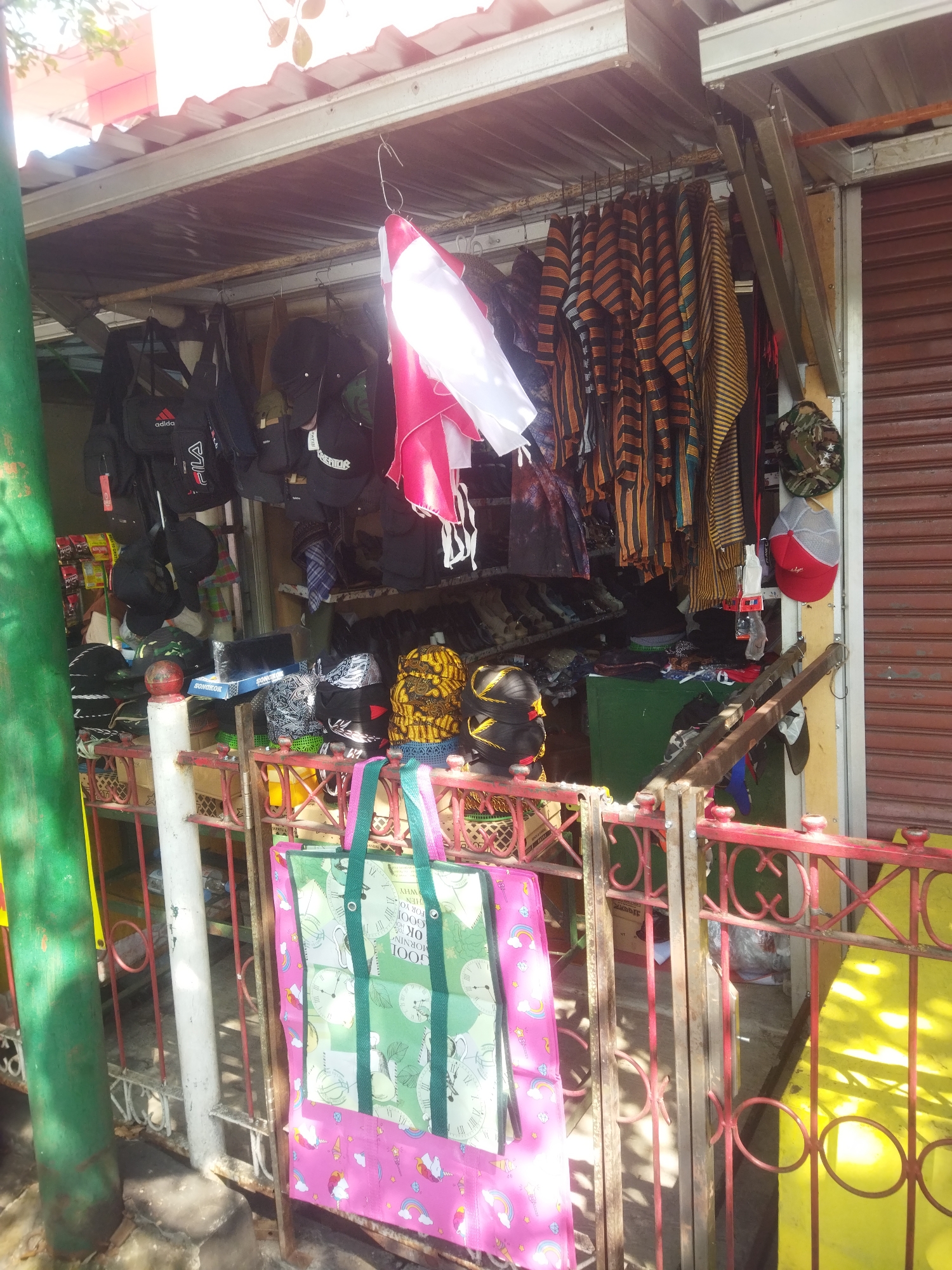 The image shows a street-side stall in Wonogiri, Indonesia, displaying various items for sale. The stall has a corrugated metal roof supported by metal beams. In the foreground, a red metal fence encloses the area. Two large tote bags, one predominantly green with clock motifs and the other pink with unicorn and rainbow designs, are hanging from the fence.
Behind the fence, the stall is filled with merchandise. Hanging from a rail are several striped shirts or robes in orange, black, and yellow. To the left, a collection of black bags, including what appears to be a backpack with "adidas" branding, are displayed. Various caps are also visible, some black, one with a camouflage pattern, and a red and grey cap.
On shelves and surfaces within the stall, more items are arranged. This includes stacks of caps, boxes, and what appear to be shoes or sandals. A green cabinet is partially visible at the back of the stall. A red and white Indonesian flag is also seen hanging. The overall impression is of a modest, open-air retail space.