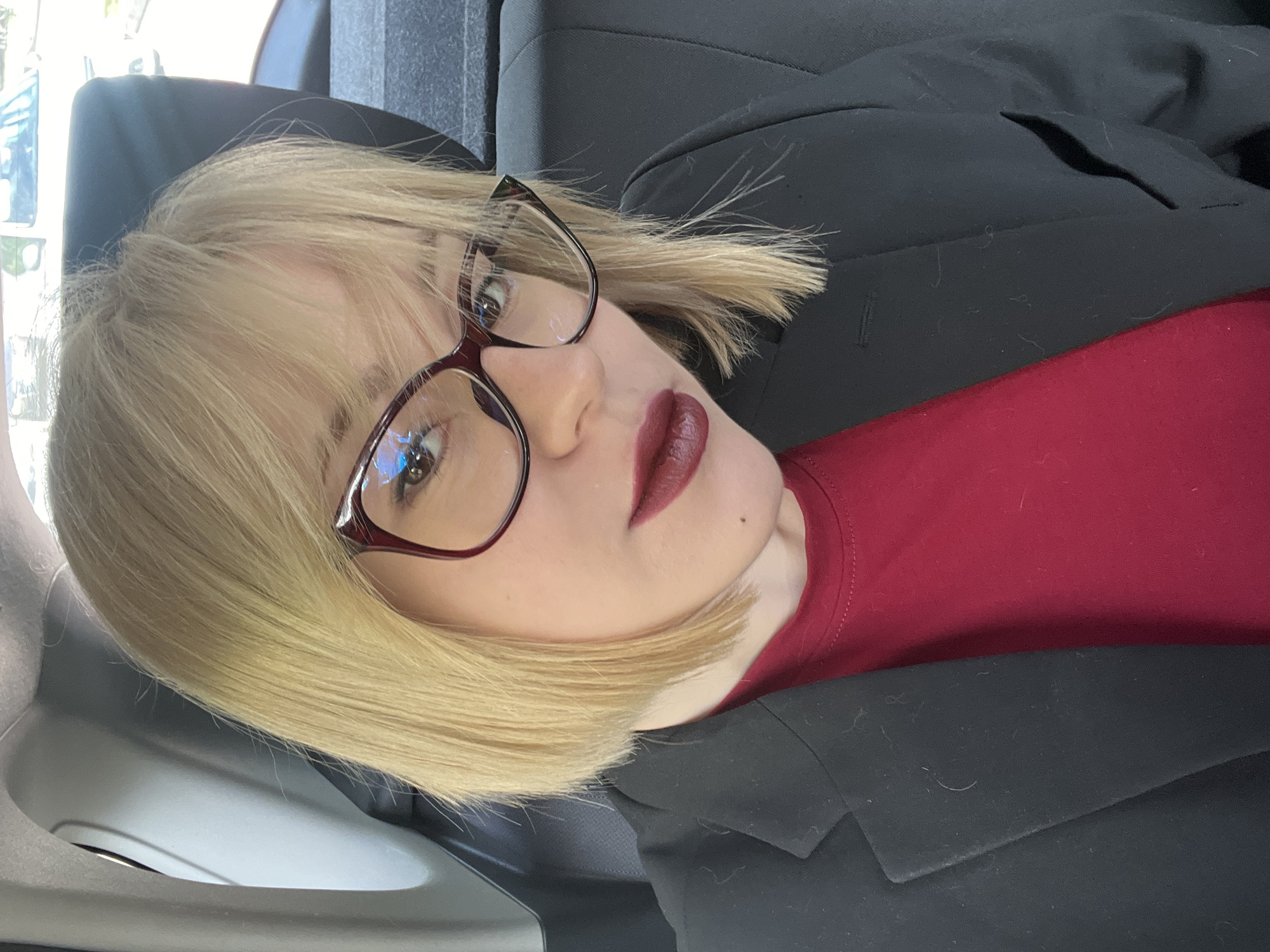 A blonde woman wearing glasses, a red top, and a black blazer is in a vehicle. The location cannot be determined as Budva, Montenegro without additional information.