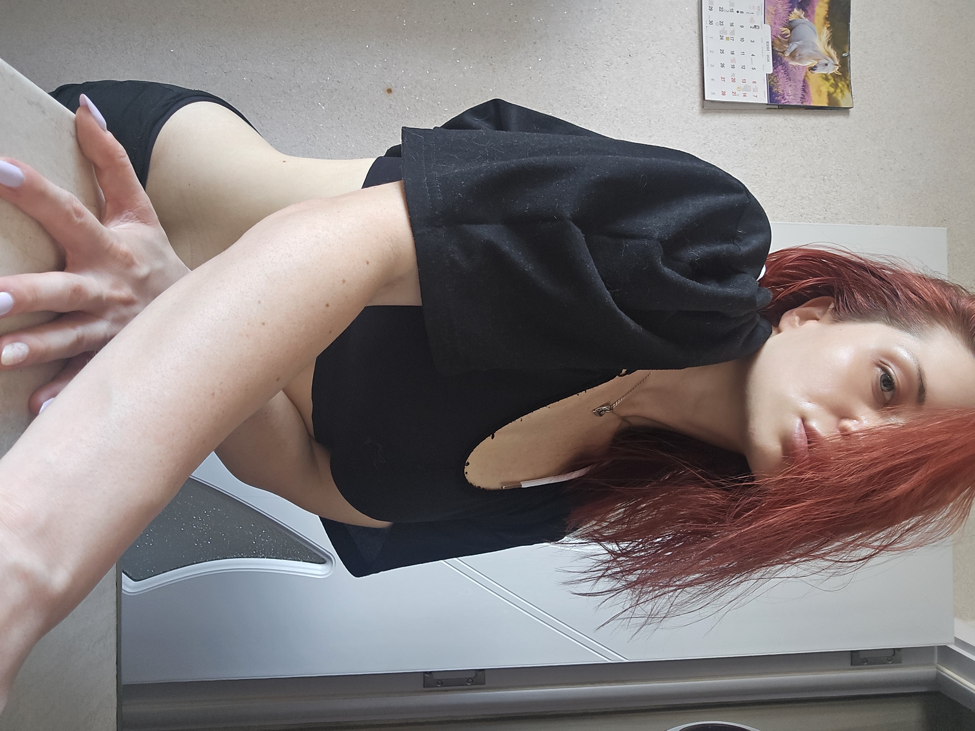 An indoor shot, rotated 90 degrees clockwise, captures a young woman with long, wavy red hair, fair skin, and light-colored eyes. She is depicted from the waist up, leaning or lying on a light-colored surface, possibly a counter or table. Her left hand, adorned with light purple nail polish, rests on this surface. She wears a black cropped long-sleeve top or hoodie over a white bra or camisole, and black bottoms. A delicate silver necklace is visible around her neck. Her head is tilted, and her vibrant red hair cascades around her face as she looks towards the camera with a relaxed expression.
The background reveals a plain, light-colored wall with a calendar hanging on it. The calendar displays "ЮНИ 2026" (JUNE 2026) and features an image of a white horse in a lavender field. Dates from 1 to 30 are visible, with 6, 7, 13, 14, 20, 21, 27, and 28 clearly readable. Below the calendar, a white piece of furniture, likely a cabinet or door, is visible. The woman's skin shows some freckles, and her face exhibits a slight sheen. The overall setting is casual and intimate, located in Burgas, Bulgaria.