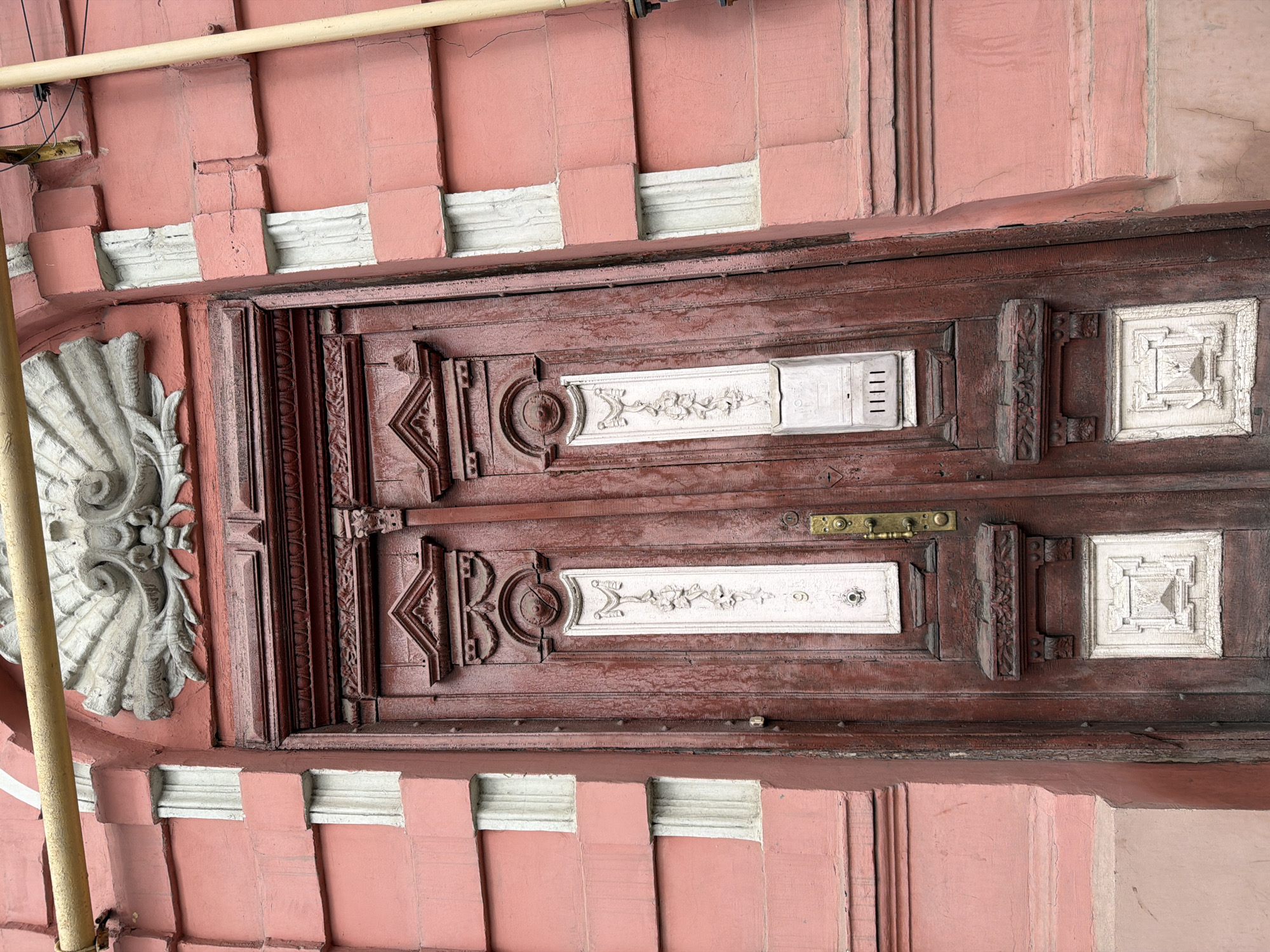 The image shows a detail of an old wooden door with decorative elements set within a pink stone frame. The door is painted a reddish-brown color and features two ornate, rectangular white panels with floral designs. There are also geometric shapes carved into the wood, including circles and triangles. The door has a brass handle and doorbell. The stone frame surrounding the door is painted pink and has a pattern of alternating rectangular blocks and white bands. A piece of scaffolding is visible on the left side of the frame. This image shows a building in Rostov-on-Don, Russia.