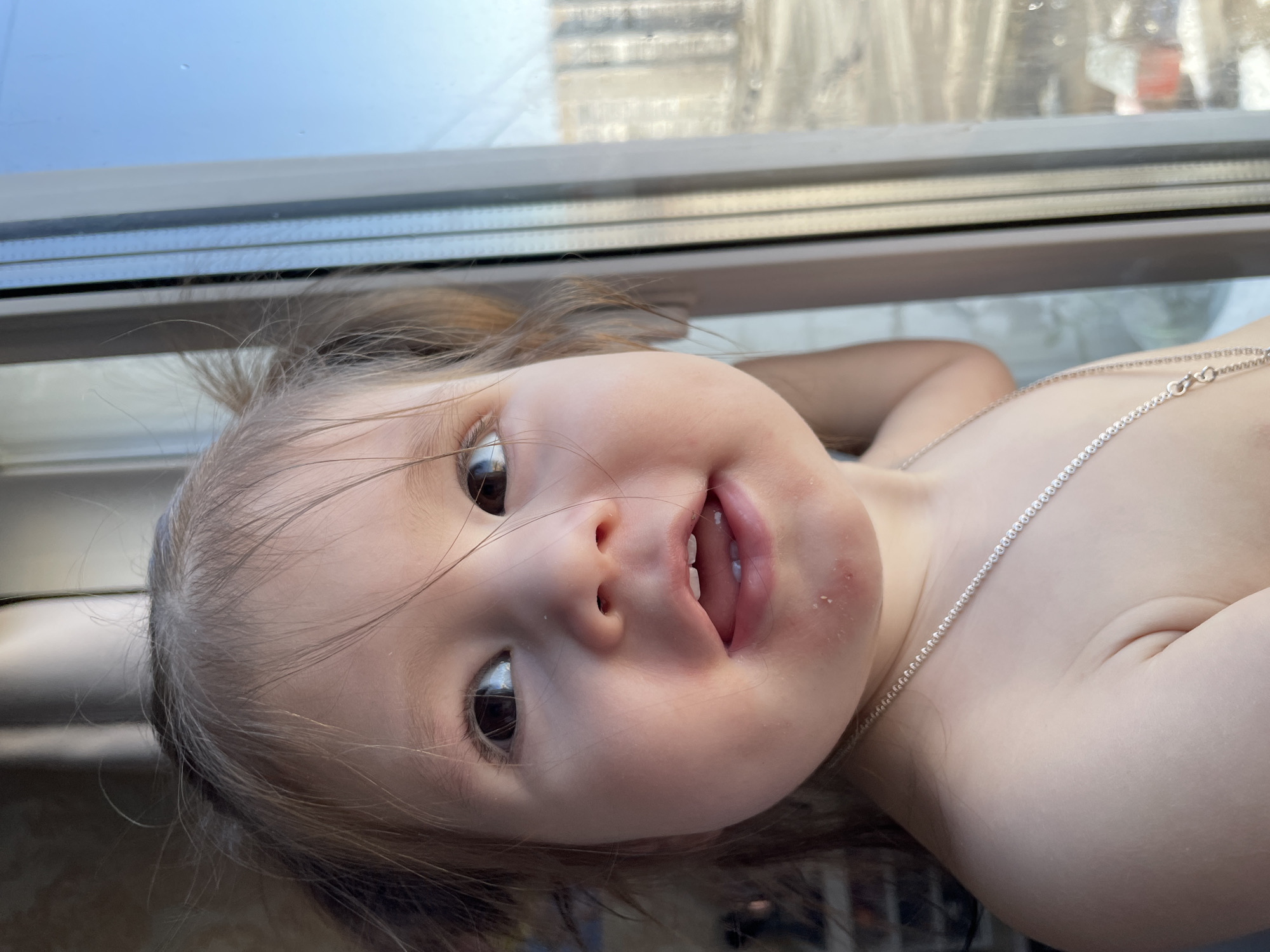 A close-up, slightly angled shot features a child with light skin, brown hair, and a silver necklace. The child is positioned near a window, with a view of a cityscape visible through the glass. The child's expression appears curious and engaging, with dark eyes and a slightly open mouth revealing a few teeth. The background shows parts of a building. The location is unspecified and cannot be identified as Izhevsk, Russia from the image alone.
