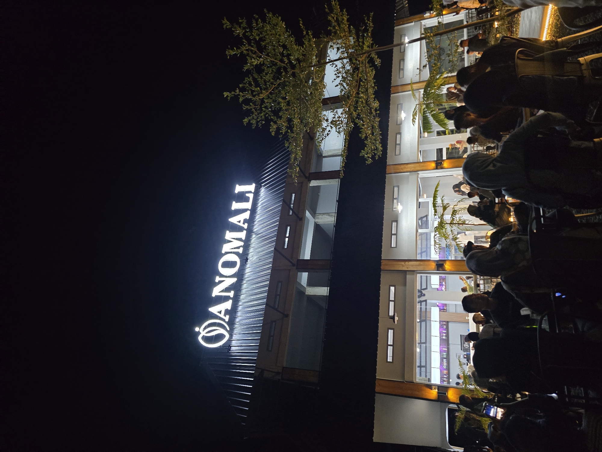 At night, in the city of Uleeue, Indonesia, a multi-story building features a prominent illuminated sign that reads "ANOMALI." The building has a dark, corrugated metal roof and a facade with large windows. Outside the building, a crowd of people are gathered, seated at tables in an outdoor seating area. The area is lit by warm-toned lights and string lights. The foreground shows silhouettes of people, with some holding up phones. In the background, inside the building, people are visible, and a bar with bottles is dimly lit. A small tree with green leaves is positioned to the right of the building. The scene depicts a lively atmosphere at an establishment, likely a restaurant or cafe, during the evening.