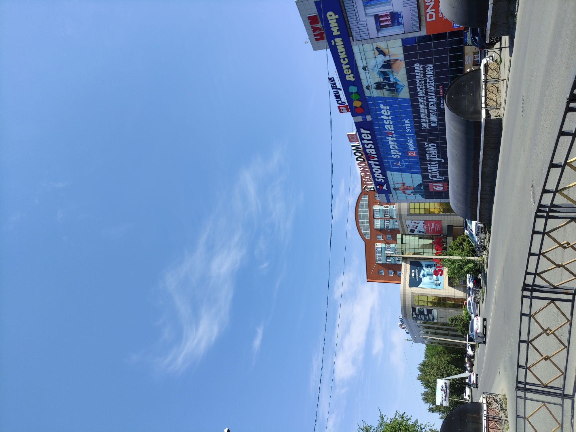 The image captures a street view in the city of Semey, Kazakhstan, on a bright, clear day. The foreground features a paved road with several cars driving or parked on the right side. A dark, arched structure, possibly a bus shelter or an underground passage entrance, is visible along the roadside. Beyond the road, a row of modern commercial buildings dominates the scene. These buildings display prominent signage for various businesses, including "TECHNODOM.KZ," "Sportmaster," "GLORIA JEANS," and "ДЕТСКИЙ МИР" (Children's World). The architecture of the buildings is contemporary, characterized by large glass facades and a mix of colorful and metallic elements. Trees and green foliage are interspersed between the buildings and along the roadside, suggesting a moderately urban environment with some greenery. The sky is a vibrant blue with thin, wispy clouds. A traffic camera is visible on a pole in the upper left portion of the image. The overall impression is one of a bustling commercial district within the city.