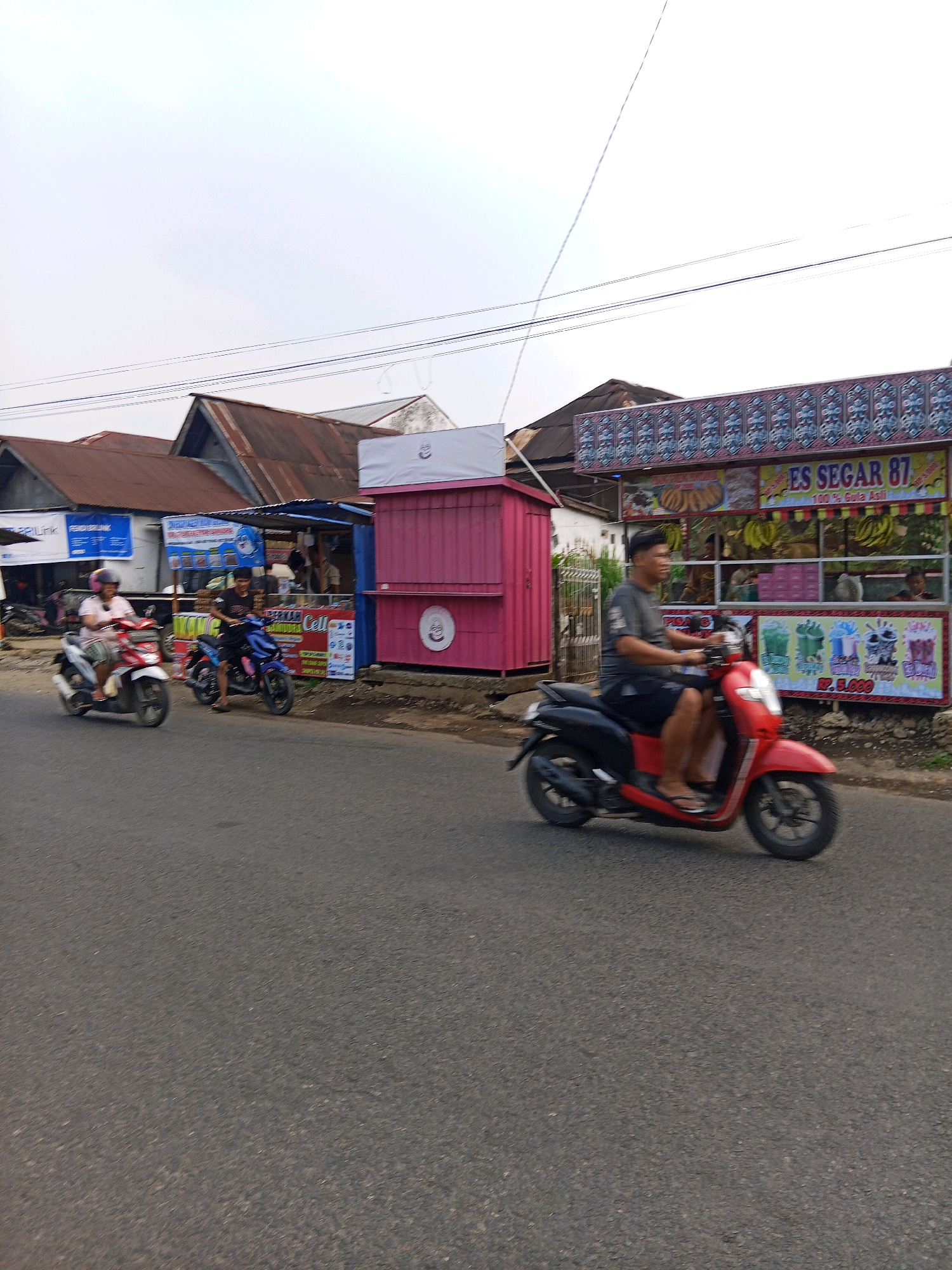 The image depicts a street scene in Lubuklinggau, Indonesia, characterized by a row of roadside stalls and shops. In the foreground, a red scooter is being ridden by a man wearing a gray t-shirt and dark shorts, with his feet out. Behind him, a blue scooter is ridden by a person in a black t-shirt and dark shorts. To the left, a woman in a pink shirt and helmet rides a white and red scooter. The stalls feature colorful signage, one prominently displaying "ES SEGAR 87" and images of fruit like bananas. Another stall has a pink facade. The background reveals buildings with corrugated metal roofs, suggesting a commercial or residential area. The road is paved, and the sky is overcast. The overall impression is of a typical street scene in a moderately busy urban area in Indonesia.