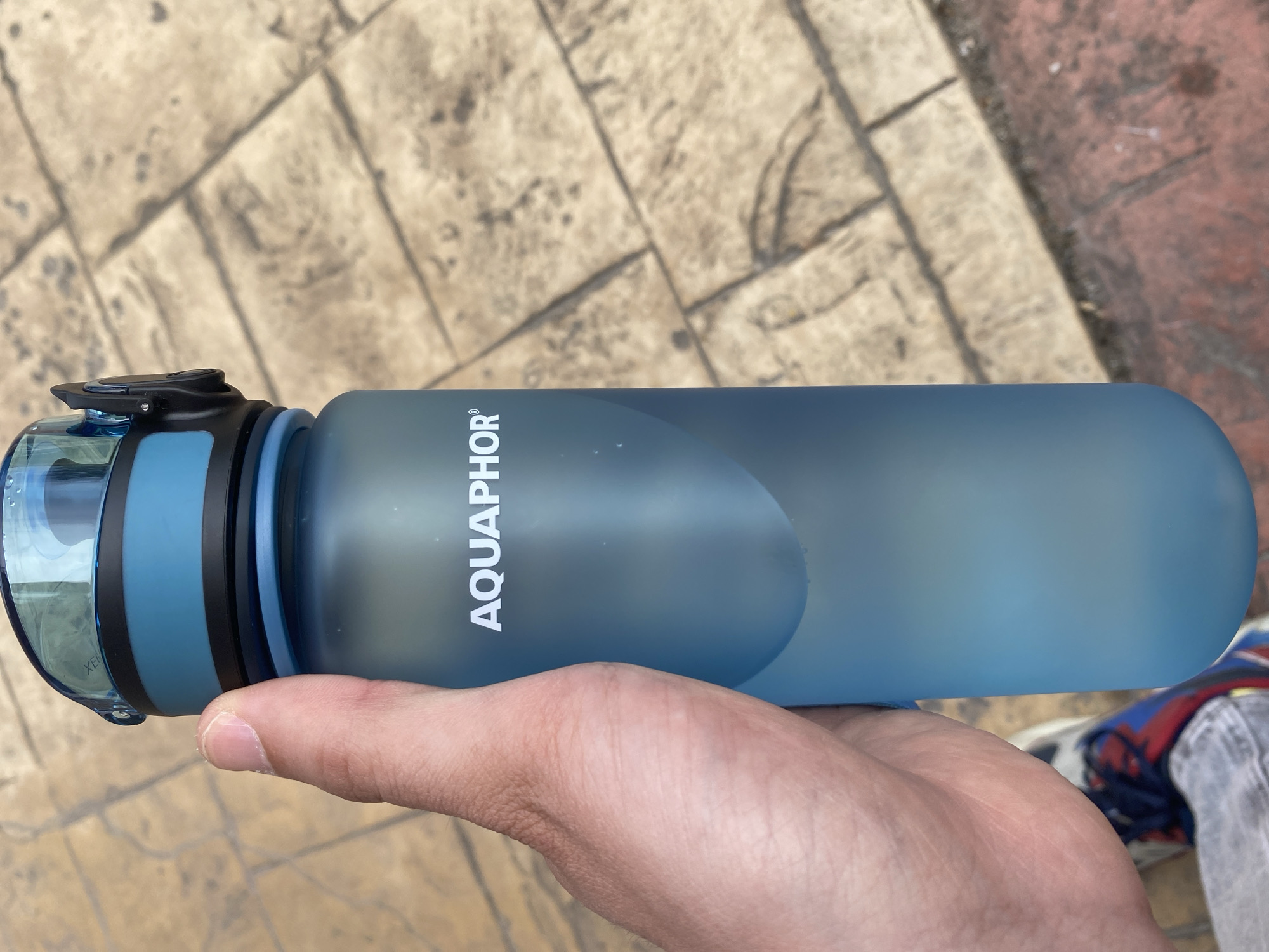 A close-up, slightly upward-angled shot shows a person's left hand holding a translucent blue "AQUAPHOR" branded water bottle. The bottle has a blue and black lid with a hinged flip-top spout. The background consists of light-colored paving stones and some reddish-brown bricks, suggesting an outdoor urban environment. The person is wearing gray jeans and a portion of colorful sneakers is visible at the bottom of the frame. The lighting indicates it is daytime, possibly overcast given the diffused light and lack of strong shadows. There are no other people or distinct landmarks to indicate a specific location within Smolyan, Bulgaria.