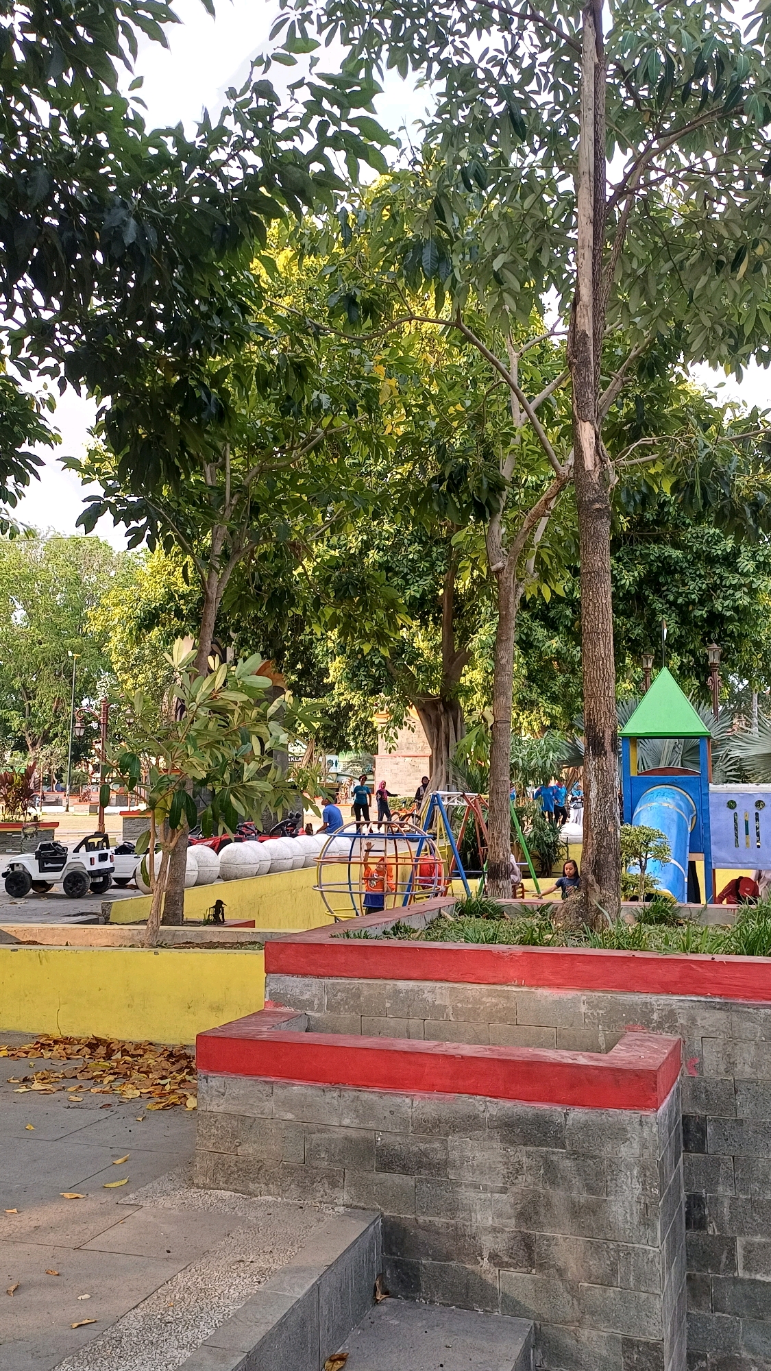 The image depicts an outdoor children's playground area situated in a park in Klaten, Indonesia. In the foreground, a tiered structure constructed with gray stone blocks and topped with red concrete beams is visible, with steps leading up from a tiled walkway. To the left, a bright yellow concrete wall borders the playground, with fallen leaves scattered on the ground in front of it.
The playground itself features a multi-colored climbing structure with a blue slide and a green roof, as well as a circular climbing frame with orange and yellow elements. Several children are present in the playground; some are playing on the equipment, while others are standing or walking nearby. A white toy car is parked to the left of the playground, near the yellow wall. The background is dominated by lush green trees, suggesting a well-maintained park environment. The overall scene captures a lively, recreational space within an urban park setting in Klaten.