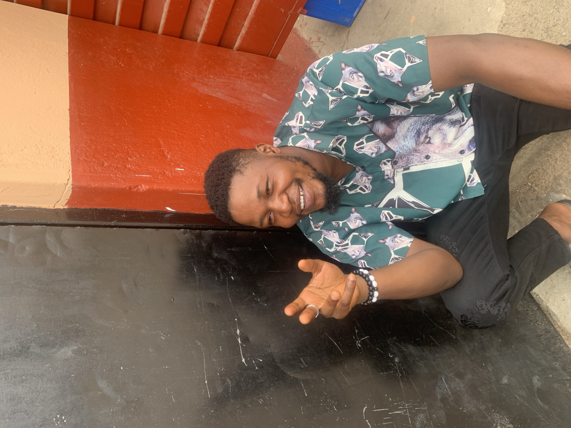 A man lies horizontally on what appears to be a paved or concrete surface. He is light-skinned, has a short haircut, a beard, and is smiling. He wears a short-sleeved shirt with a pattern of wolf faces, black pants, and a beaded bracelet on his left wrist. He extends his right hand towards the center of the frame. The man is lying near a wall, which has a yellow and red painted upper portion and a black painted lower portion. The location is suspected to be Jos, Nigeria.