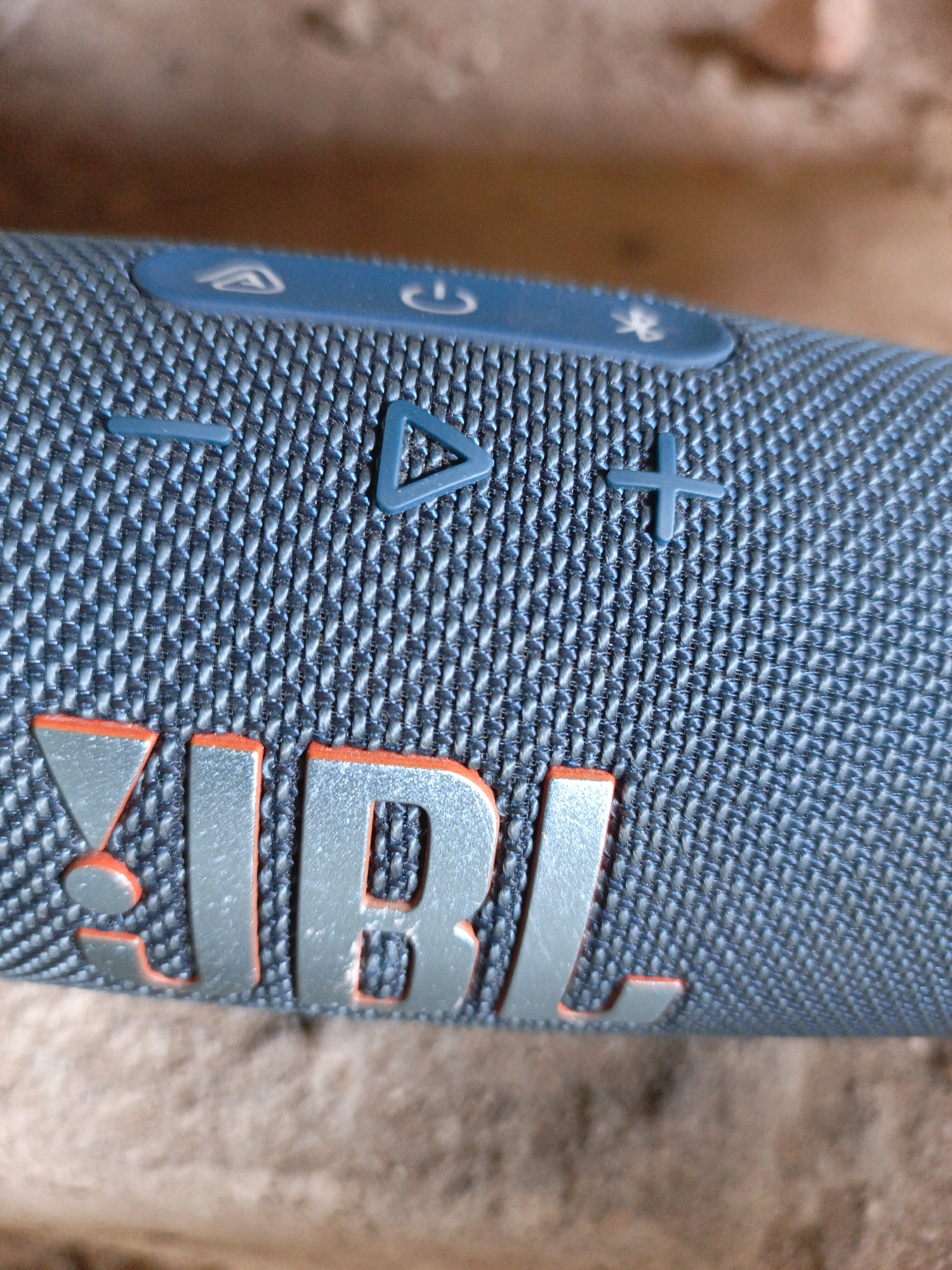 Close-up shot of a blue JBL speaker on a brick surface. The JBL logo is in silver with an orange outline. Above the logo are the speaker's control buttons, including volume controls, a play/pause button, and a power button. The speaker's fabric has a woven texture. The location is unknown.