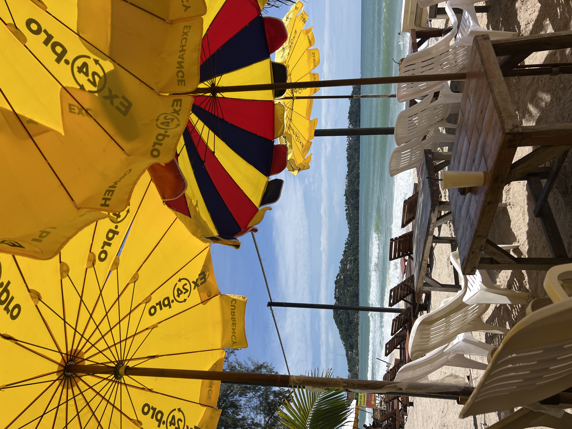 The image depicts a sandy beach scene on a clear, sunny day. The foreground is dominated by the undersides of several large beach umbrellas, primarily yellow. The yellow umbrellas display reversed text and a currency exchange symbol, with "EXCHANGE" and "CURRENCY" legible in some instances. To the right, a multi-colored umbrella with red, yellow, and blue striped panels is visible. Beneath these umbrellas, multiple wooden tables and white slatted plastic chairs are arranged directly on the sand. A yellow cup with a straw rests on one of the tables in the lower-middle right.
In the midground, the sandy beach extends towards the ocean, where several wooden beach loungers are visible. The ocean features light-to-moderate waves breaking near the shore. A person is minimally visible swimming in the ocean on the far right. A partially obscured sign on the left mentions "Eats and Cannabis." Palm foliage is visible on the far left. The background features a large, green, hilly landmass covered with vegetation and some structures, likely an island or distant coastline, under a bright blue sky with scattered white clouds. The location is a beach, specifically an outdoor cafe or relaxation area.