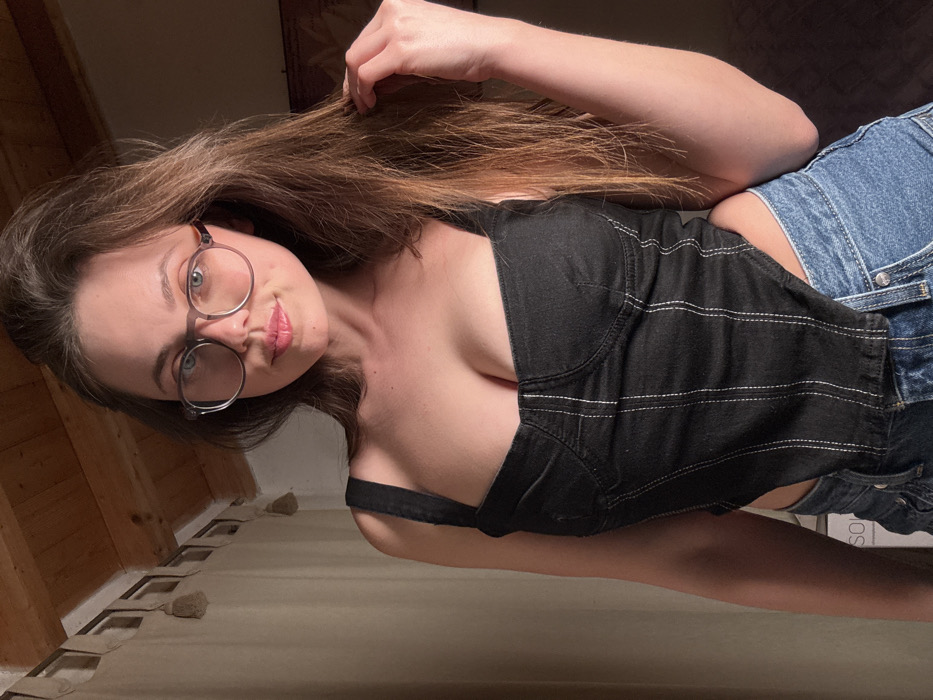 A forensic examination of the image reveals a single female subject captured in a posed, mid-shot composition within an indoor setting, identified by contextual information as a location in Monsummano Terme, Italy.
The subject is a young adult female with light skin and long, brown, slightly wavy hair that falls over her shoulders. She wears translucent, round-framed eyeglasses with clear lenses. Her facial expression is neutral, with her gaze directed forward, mouth slightly open. She is dressed in a black, form-fitting, sleeveless crop top with wide shoulder straps, featuring a structured bustier design and prominent vertical white stitching on the front. The bottom hem of the top is asymmetrical, revealing a section of her left midriff. Below the crop top, she wears blue denim jeans, partially visible. Her left hand is raised, with fingers positioned within her hair on the right side of her head.
The immediate background to the subject's left consists of a light-colored, possibly beige, curtain or fabric wall covering. Above, exposed wooden ceiling beams are visible. To the subject's right, the background is darker and less defined, suggesting a further recessed area of the room or blurred furniture. Illumination appears to be internal and warm, casting soft light on the subject.