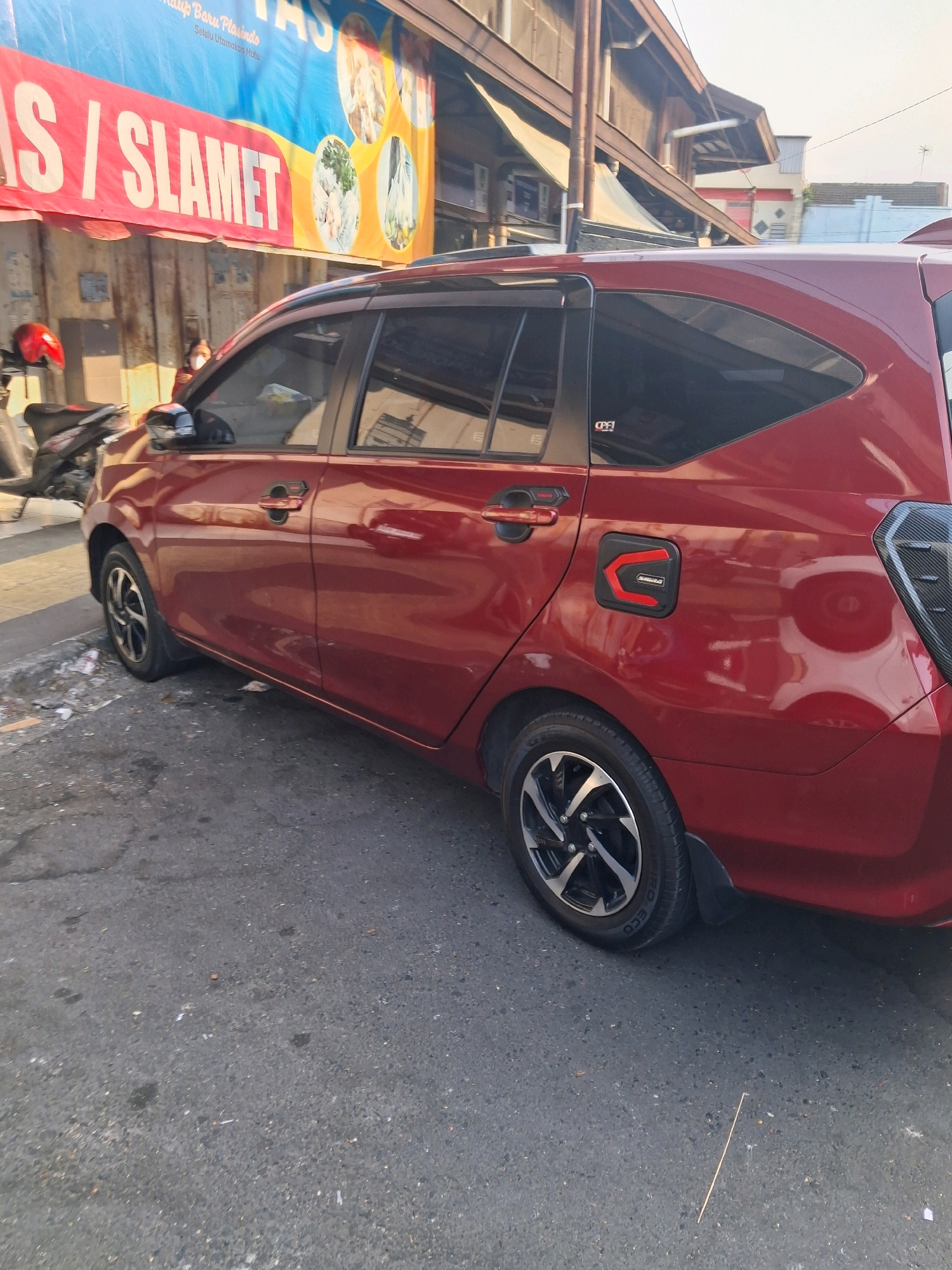 A red car is parked on a paved street in front of a building with a banner advertising "TAS" and "SLAMET" in Surakarta, Indonesia. A motorcycle is parked to the left of the car. The car has tinted windows and a black fuel cap with a red accent. The street is dirty with some debris visible. The building in the background appears to be a shop or business. The sky is bright, suggesting it is daytime.