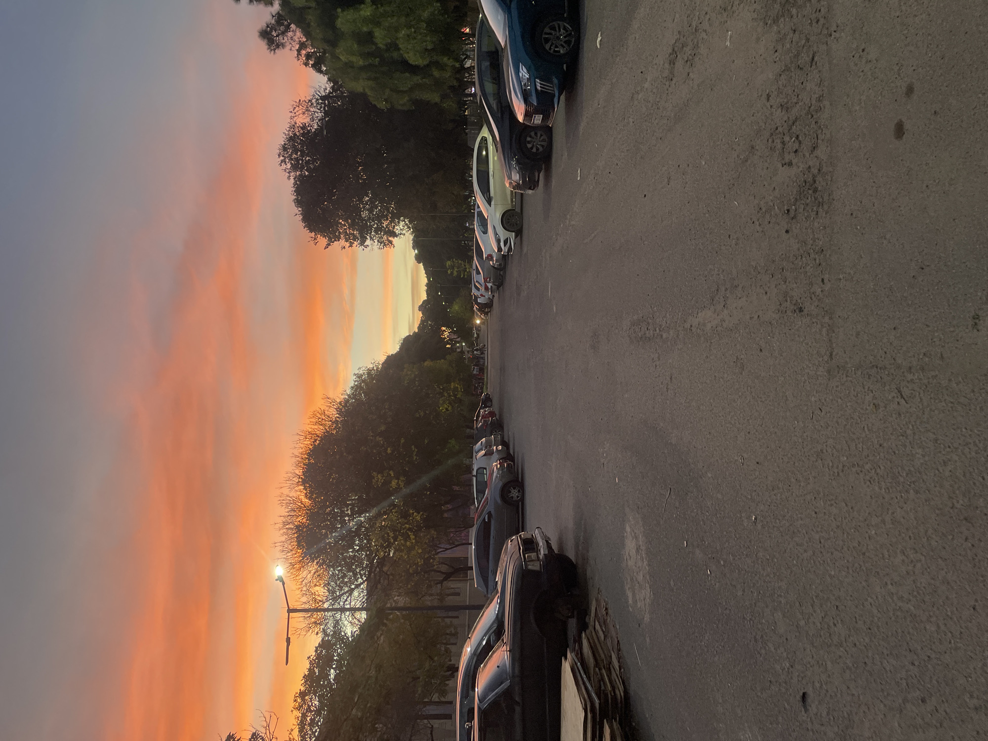 The image shows a street in Buenos Aires, Argentina, at dusk. Cars are parked along the right side of the street, and trees line the left side. The sky is a mix of orange, yellow, and grey. A street lamp is visible on the left.