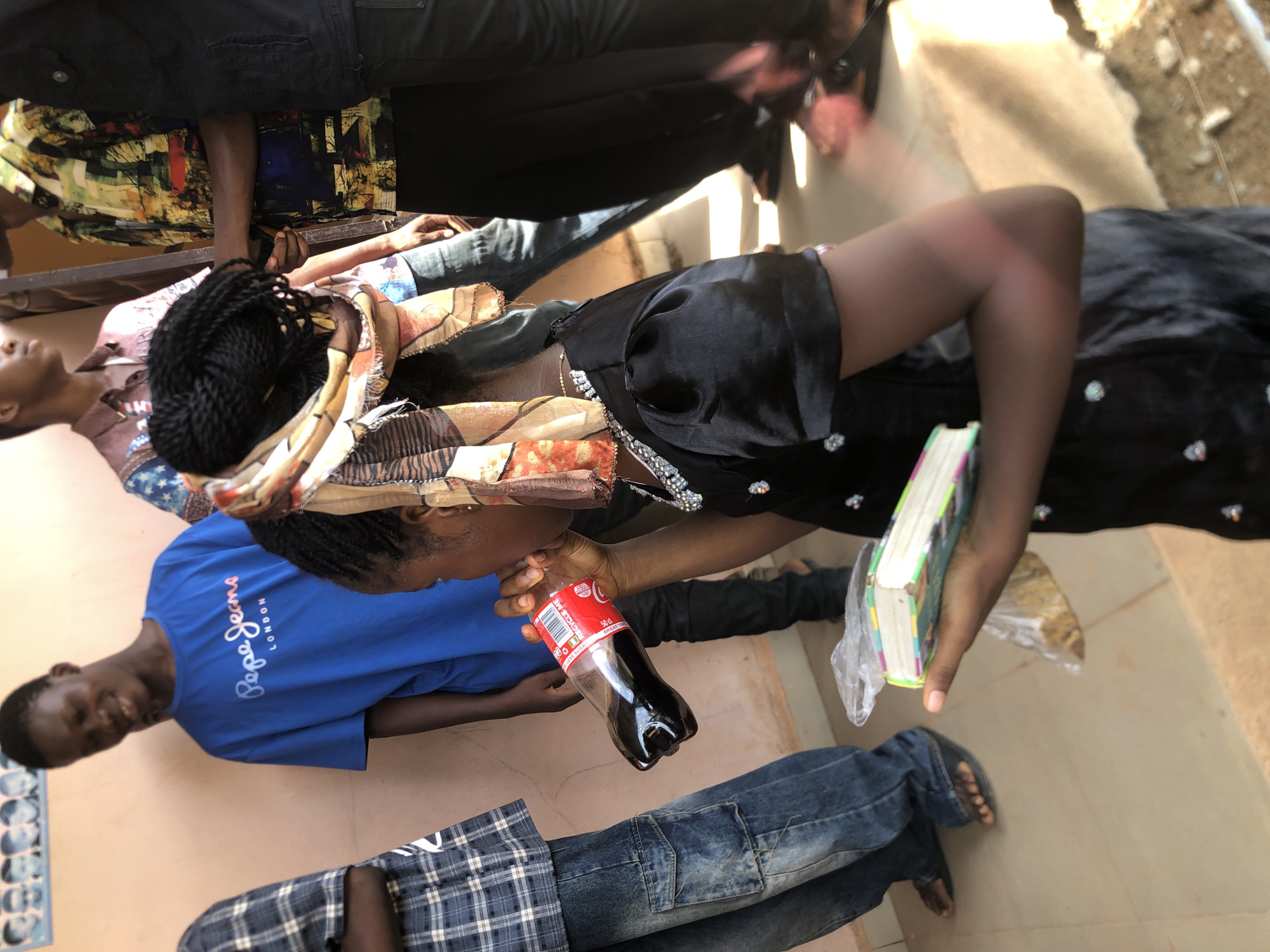 In Nkwelle Ezunaka, Nigeria, the image shows a woman drinking from a Coca-Cola bottle. She is wearing a black dress with embellishments and a patterned headscarf. A book wrapped in plastic is in her other hand. A person in a blue "Pepe Jeans LONDON" t-shirt is nearby, and another person is visible in a plaid shirt and jeans. Other individuals are partially visible, including one in a brightly colored shirt and another in black pants and a dark top. The location seems to be indoors with tiled flooring. A poster is partially seen on the wall to the left. Outside, there appears to be dirt and debris near a wall.