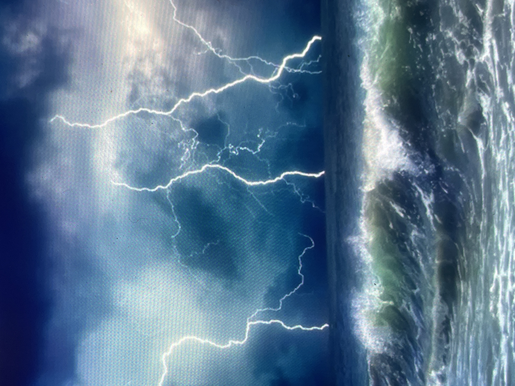 The image, contextualized as being located in the city of Zelenyi Hai, Ukraine, depicts a severe marine thunderstorm over a turbulent ocean. The sky is dark and heavily overcast with dense, ominous blue-grey clouds. Multiple bright white lightning bolts streak vertically from the clouds towards the ocean surface; at least four distinct strikes are visible, with some exhibiting jagged, forked paths. The horizon line is indistinct due to the dark conditions and rough sea. The ocean below is agitated, characterized by large, breaking waves with significant white foam and spray. The water appears in shades of deep blue and greenish-grey. No people, vessels, or landmasses other than the ocean itself are visible in the scene.