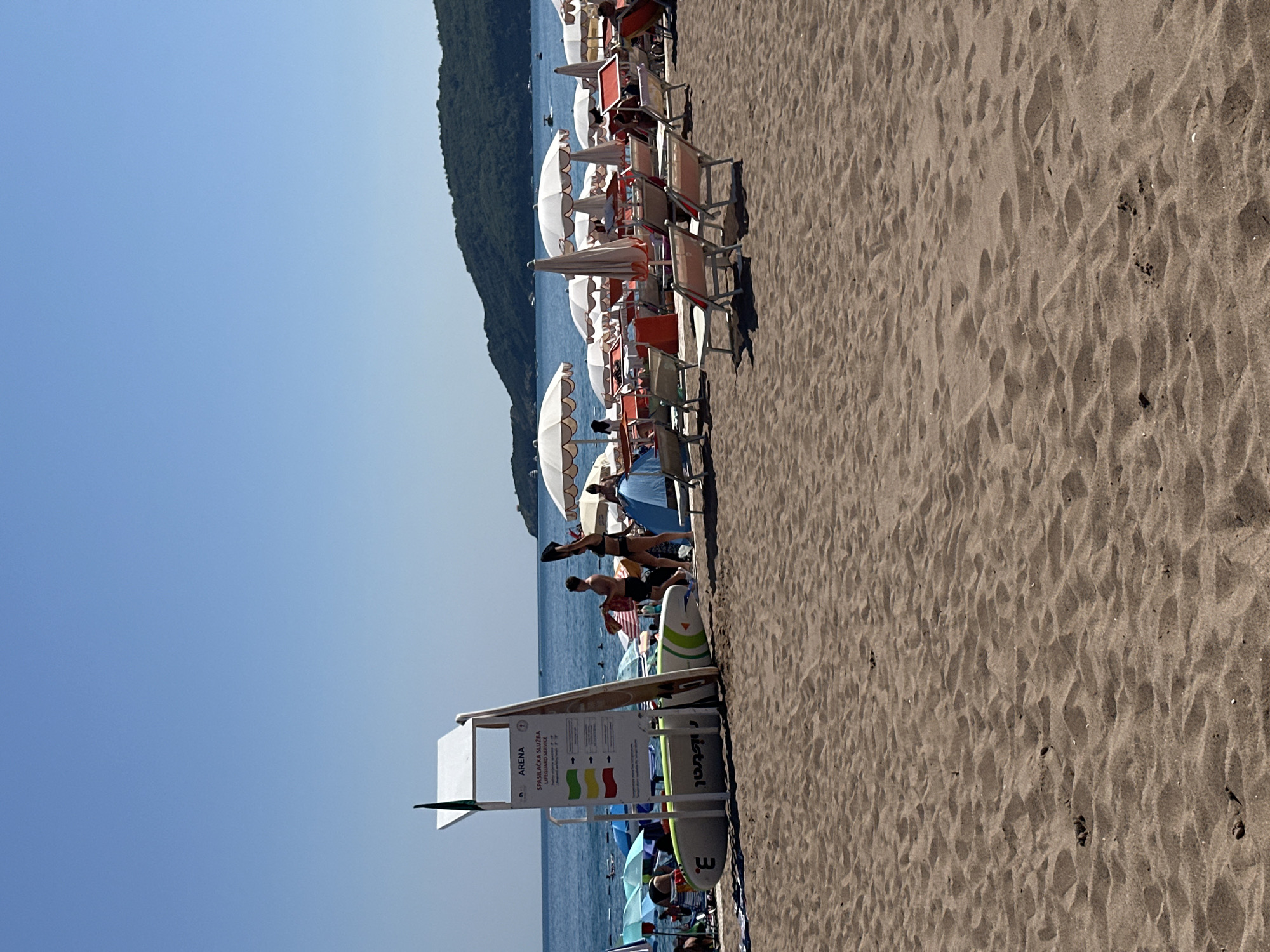 This is a vertical, medium-shot photograph of a crowded beach on a sunny day. The sand in the foreground is marked with footprints and ripples. Rows of white beach umbrellas and lounge chairs are set up on the sand, some occupied by people. The sea is visible in the background, with a forested hill rising steeply from the water's edge. The sky is a clear, bright blue.
Several people are visible on the beach, some lounging in chairs or under umbrellas, and others standing or walking. Two individuals are particularly noticeable: a man in black shorts doing a plank exercise on the sand, and another man in the background standing in the water. The scene appears to be a popular tourist destination, likely during the summer season, given the number of people and the bright, sunny weather. The overall mood is relaxed and recreational.
A signpost is visible on the left side of the image, displaying information in what appears to be Croatian or a similar Slavic language. The text reads "ARENA SPASILAČKA SLUŽBA LIFEGUARD SERVICE" along with colored flags indicating water safety conditions. Other objects on the beach include surfboards and what appear to be paddleboards. The image captures a typical summer beach scene with ample sunlight and a clear, cloudless sky, suggesting it was taken during the daytime, likely mid-morning or afternoon. The location is Boreti, Montenegro, as indicated by the context.