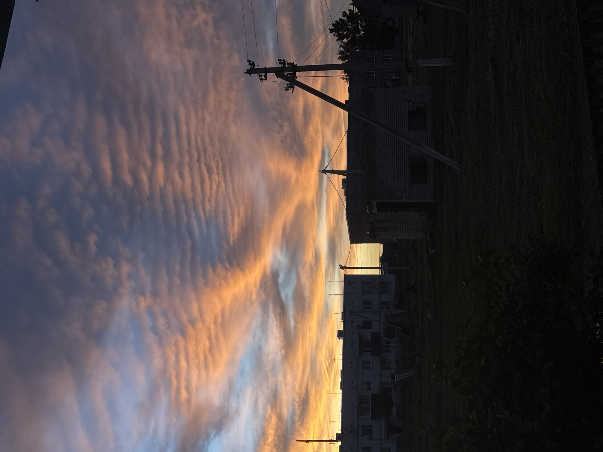 This image depicts a dusk sky over the city of Mursalimkino, Russia. The sky is filled with dramatic clouds painted in shades of orange, pink, and blue, with the sun's warm glow illuminating the horizon. In the foreground, silhouetted against the vibrant sky, are several buildings. To the left, a multi-story apartment building stands, its windows dark. To the right, a smaller, unfinished brick building is visible, with visible beams supporting its structure. Power lines and poles crisscross the scene, adding a utilitarian element to the landscape. The ground is covered in grass, and a dark, shadowy area in the immediate foreground suggests dense foliage or a shaded balcony. The overall impression is one of a peaceful, yet slightly desolate, urban twilight.