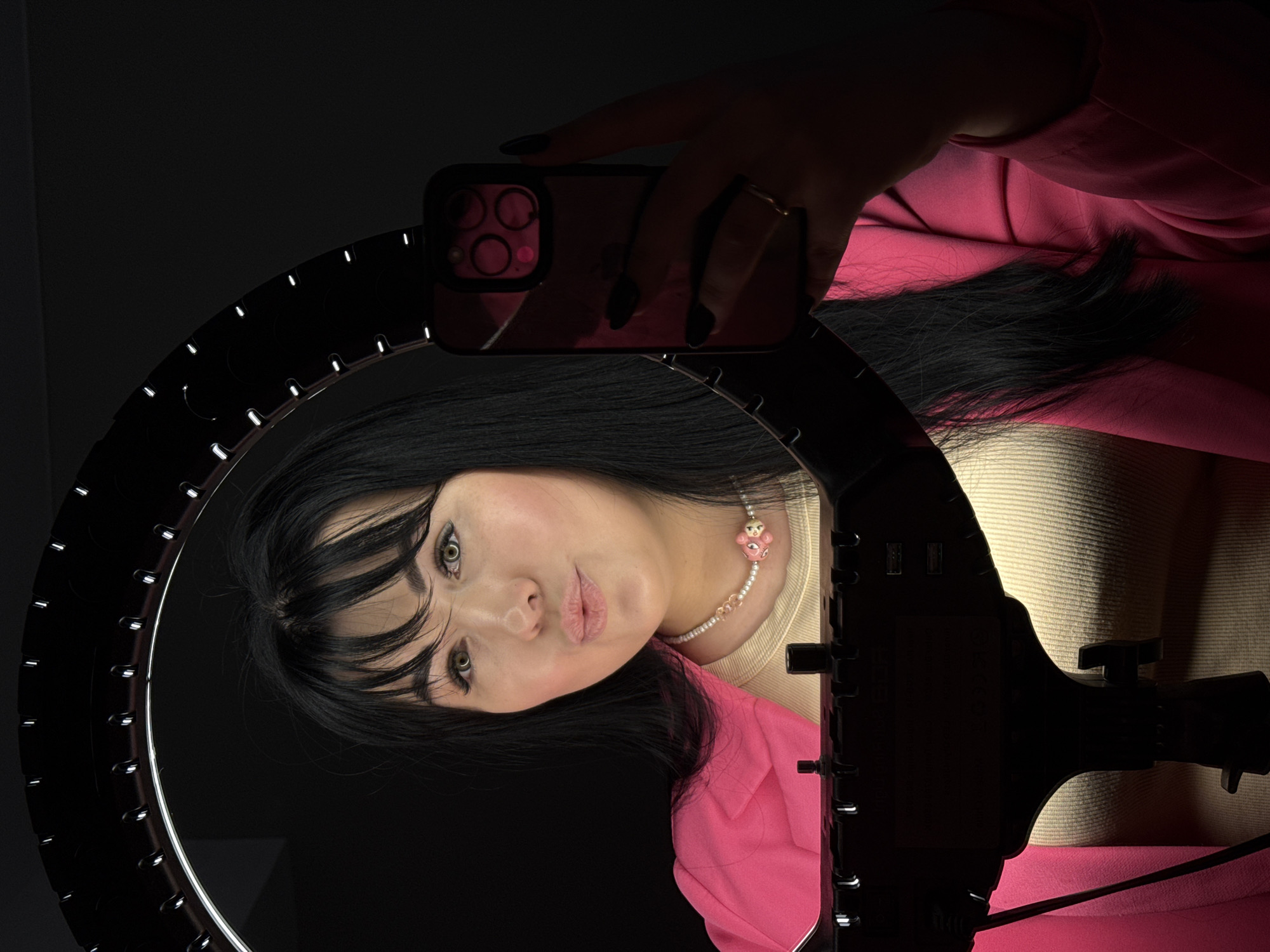 A woman with black hair and bangs, wearing a pink blazer, is holding a smartphone in front of a ring light. The phone has a pink case and multiple camera lenses. She is also wearing a necklace with pink bear beads. The environment appears to be indoors, with a dark background outside of the ring light's illumination. There is no information available to confirm the image was taken in Chaikovsky, Russia.