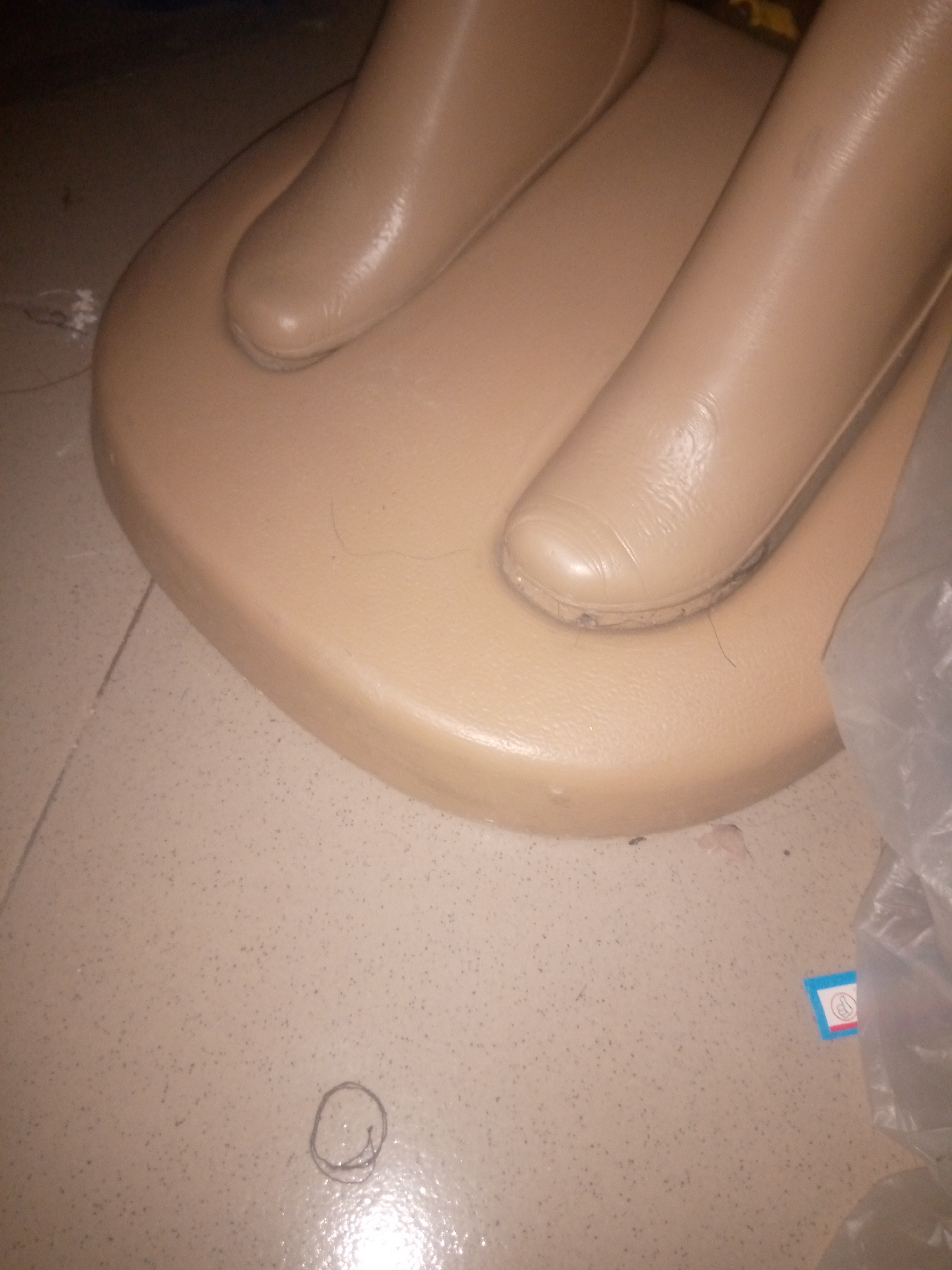 The image shows the lower portion of a mannequin, specifically its legs and feet, resting on a beige, oval-shaped base. The mannequin's legs are a pale, skin-like color and appear to be made of plastic or a similar synthetic material. They are positioned as if standing on the base. The base itself is smooth and slightly reflective, resting on a tiled floor. The floor tiles are light brown with darker speckles. There is a faint circular doodle on the floor to the left of the base. A small, blue and white sticker with an icon on it is visible on the floor to the right of the base. A crumpled plastic bag is partially visible on the right side of the image. The overall environment appears to be indoors, possibly a retail space or a storage area. There are no people visible in the image, and no discernible location markers place this scene within Jalingo, Nigeria.