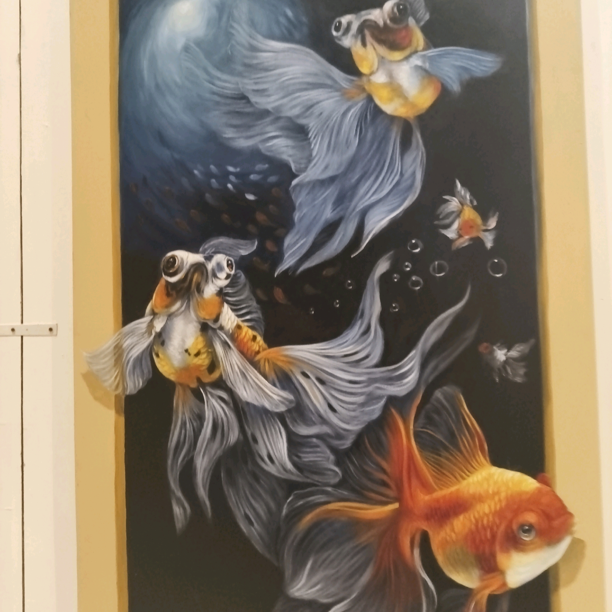 This is a vertical, full-shot, eye-level, medium-light outdoor photograph of a framed painting of goldfish. The painting depicts several goldfish with flowing fins against a dark background, illuminated by a bright, swirling light source from the upper left. The painting is framed by a yellow border and is mounted on a light-colored wall. The surrounding environment suggests an indoor setting, likely an art gallery or home, with a portion of a white door visible on the left side. The goldfish are rendered with fine detail, emphasizing their scales and the translucent quality of their fins. Bubbles are scattered throughout the painting, adding to the underwater illusion. The painting's style is realistic with a touch of surrealism due to the dramatic lighting and exaggerated fins. No people are visible in the photograph.