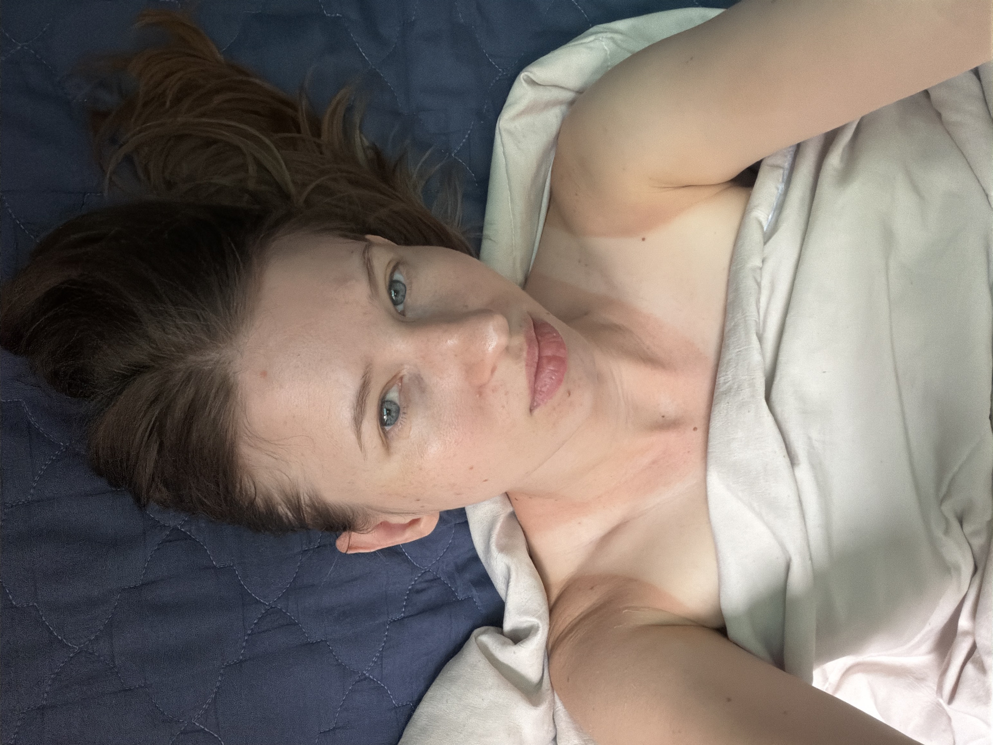 An overhead, close-up shot captures a fair-skinned woman lying on her back on a dark blue quilted comforter in Dongducheon-si, South Korea. Her head is positioned at the top of the frame, with her long, light brown hair fanned out across the comforter. She is wearing a light-colored sheet or blanket that covers her chest and abdomen, exposing her shoulders and upper arms. The woman's eyes are open and looking directly at the camera, with a neutral expression. Her skin shows distinct tan lines on her shoulders and neck, indicating recent sun exposure. The lighting suggests an indoor setting, possibly a bedroom.