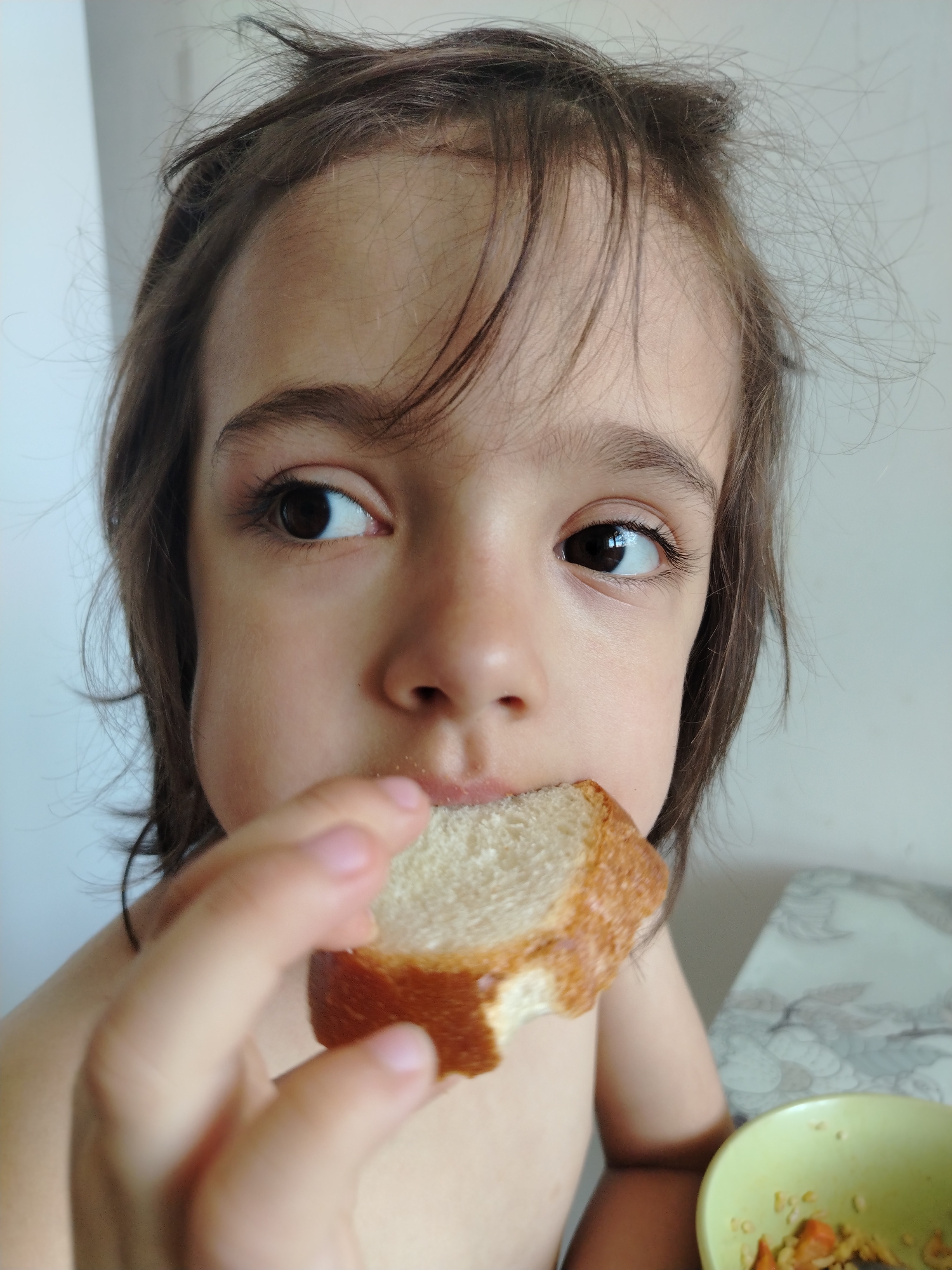 A close-up shot shows a child with brown hair and eyes eating a piece of bread. The child is holding the bread with their right hand, with a visible bite taken out of it. A green bowl containing food is visible in the lower right corner of the frame. The background appears to be a wall with a floral-patterned surface. The location is assumed from the given prompt to be in Noviy, Russia. The subject is a child eating.