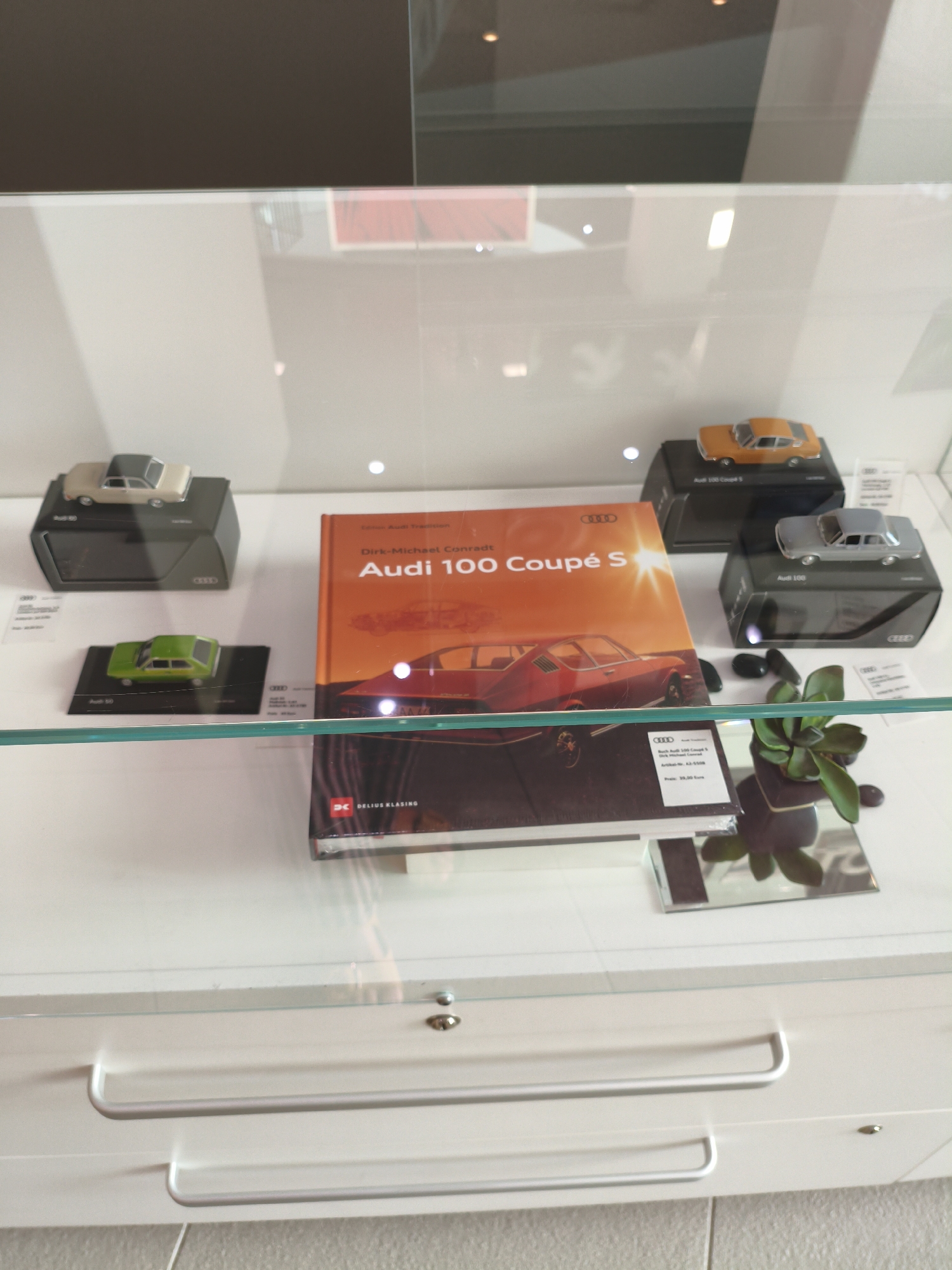 The image shows a display case in Ingolstadt, Germany, possibly in a museum or showroom. The display case contains several items, including model cars and a book. In the foreground, there is a green model car on a black stand. To the right of the green car, there is an orange book with the title "Audi 100 Coupé S" and "Dirk-Michael Conradt" written on it. Behind the book, there is a small potted plant. In the background, there are two more model cars, one orange and one silver, each on a black stand. The display case has glass doors, and reflections are visible on the glass. The overall impression is of a curated exhibition, likely related to Audi automobiles, given the presence of Audi branding on some items. The location in Ingolstadt is strongly suggested by the presence of Audi branding and the city's association with the automobile manufacturer.