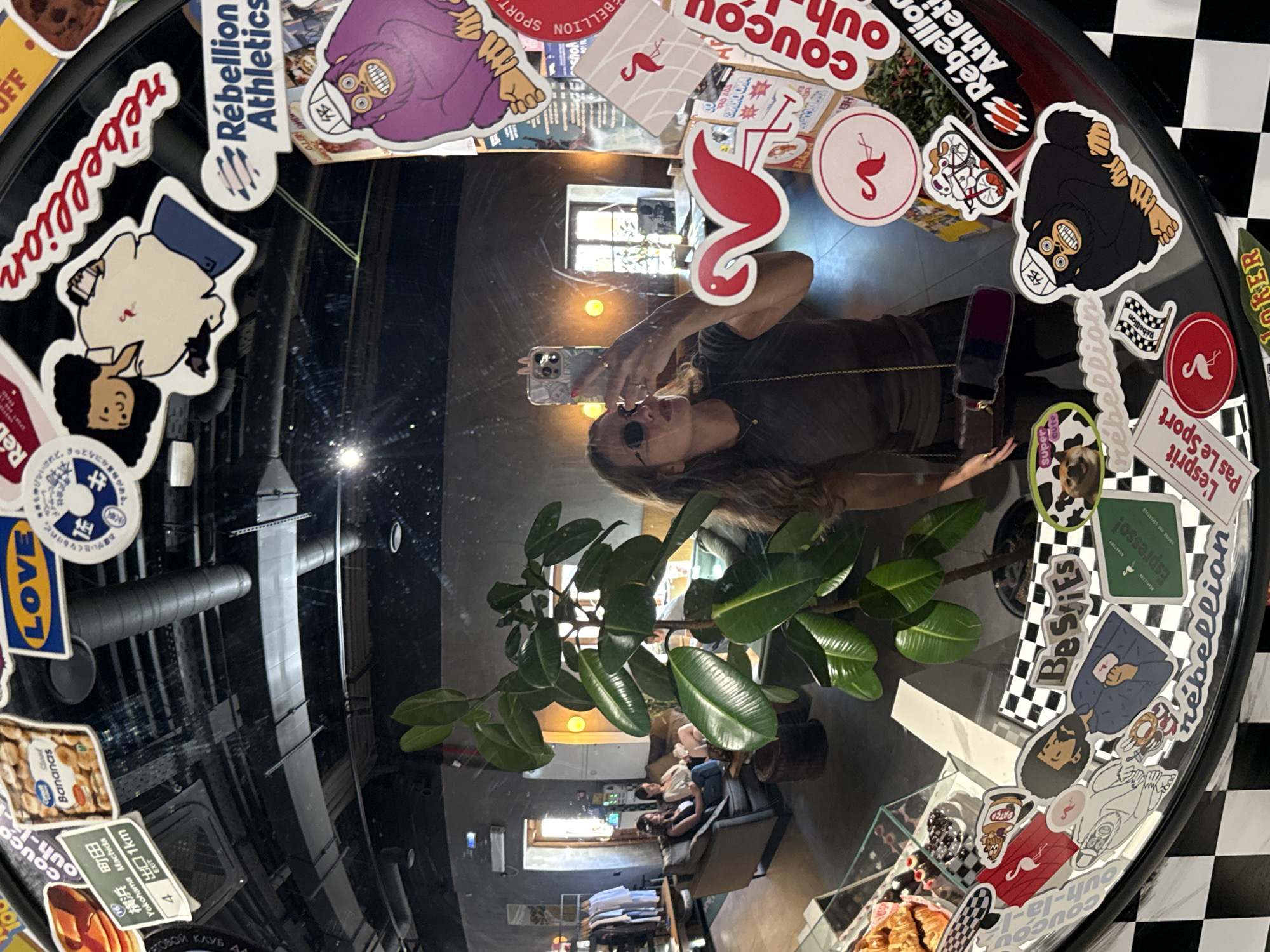 This is a photograph taken in what appears to be a cafe or a similar establishment, reflected in a circular mirror. The primary subject is a person taking a selfie in the mirror, holding up a phone. The person is wearing sunglasses and has long, brown hair. The reflection also shows a large potted plant with glossy green leaves in the foreground.
The mirror's surface is heavily decorated with various stickers, most of which are colorful and feature cartoonish illustrations, text, and logos. Some of the visible text on the stickers includes "Rebellion Athletics," "COUCOU," "LOV," "Bananas," "Espresso!," and Japanese characters that appear to be place names like "Yokohama Machida." There are also stickers depicting characters, animals, and graphic designs like flamingos and checkered patterns.
In the background, visible through the mirror's reflection, is the interior of the cafe. It seems to be daytime, with light coming from a window. There are other patrons sitting at tables and a counter area. The overall atmosphere suggests a casual and possibly trendy setting. The presence of a checkerboard pattern on some stickers and in the background hints at a possible theme.