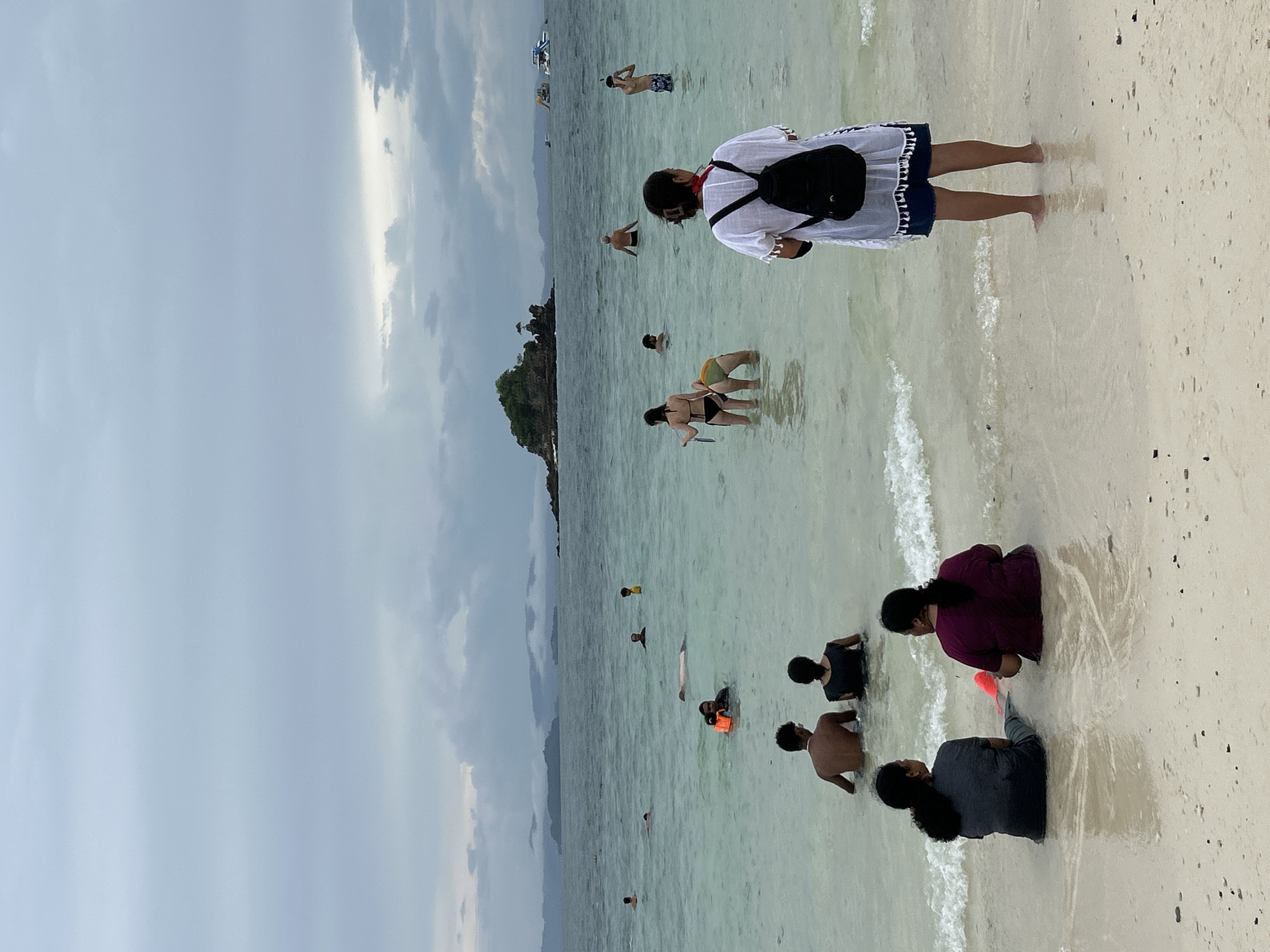The image depicts a beach scene in Phru Nai, Thailand, on an overcast day. The water is clear and shallow, with several people wading and swimming. In the foreground, two individuals are seated facing away from the camera, with their backs to the sea. To the right, another person stands facing the water, wearing a white cover-up, dark shorts, and a black backpack. In the middle ground, various people are visible in the water, some with snorkeling gear. Further out, a small, rocky island with vegetation rises from the sea, and in the background, distant islands and boats are discernible. The sky is filled with grey clouds, suggesting possibly impending rain or a recent shower. The beach itself is sandy with scattered small dark objects.