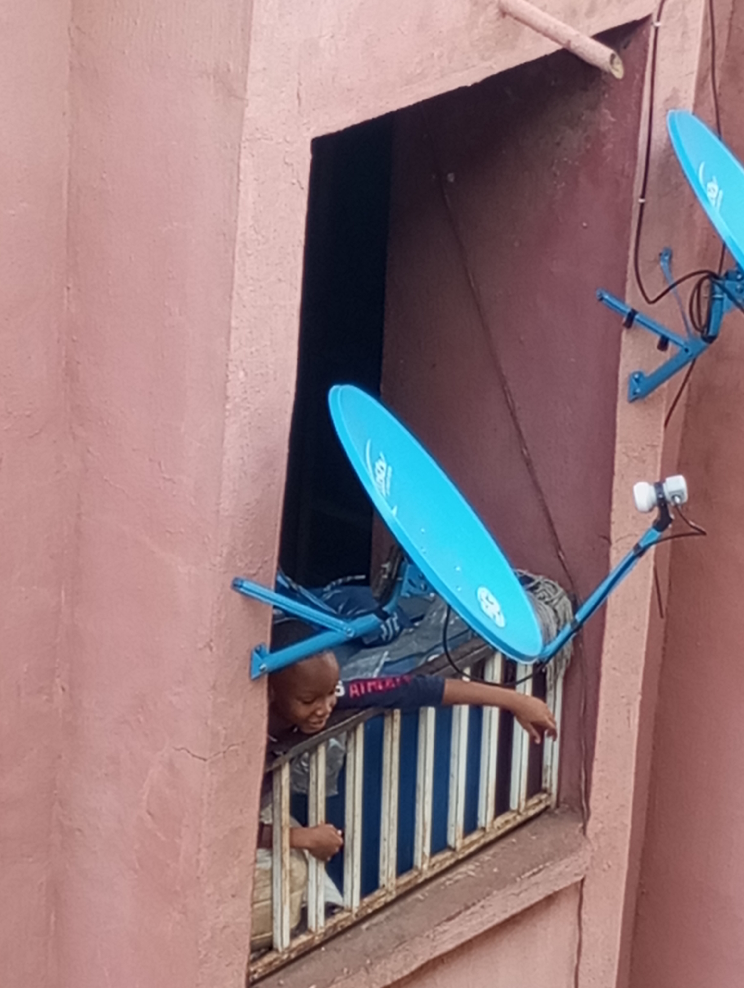 In Onitsha, Nigeria, a young boy leans out of a window, his face visible as he looks down and to his right. The boy is wearing a dark long-sleeved shirt with some lettering on the sleeve, and light-colored pants. His left arm rests on the window's railing, and his right hand is extended outward. Two bright blue satellite dishes are mounted on the exterior of the building, one directly above and to the right of the window, and another partially visible in the upper right corner of the image. The building's exterior wall is painted a reddish-brown, with a lighter pinkish-brown wall on the left side of the frame. The window is covered by a metal grate.