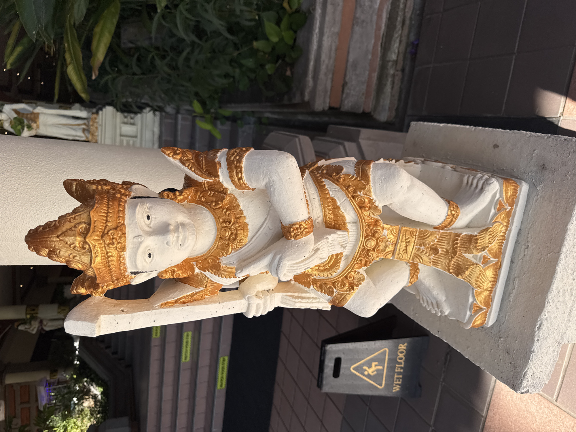 A white and gold statue of a figure, likely a deity or mythical being, is centrally positioned on a rough concrete plinth. The statue is painted white with intricate gold detailing on its crown, jewelry, and attire, depicting a crouched or kneeling posture while holding a long, flat object, possibly a weapon or staff, with its left hand. Its right hand rests on its knee. The scene is located in Kuta Selatan, Indonesia. To the left of the statue, a black A-frame "WET FLOOR" sign with a yellow warning symbol is visible on a tiled floor. Beyond the sign, a wide set of multi-tiered stairs ascends, featuring several yellow "WATCH YOUR STEP" stickers on the step edges. Further back on the left, lush green foliage is present, and a building interior with blurred details and light sources is visible at the top of the stairs. To the right of the main statue, a thick white pillar or wall stands, behind which dense green vegetation is visible. Partially obscured by the pillar and foliage, another white standing statue, also appearing to be a deity with golden accents, is visible in the background. The scene is brightly lit from the left, casting distinct shadows on the ground and parts of the main statue.